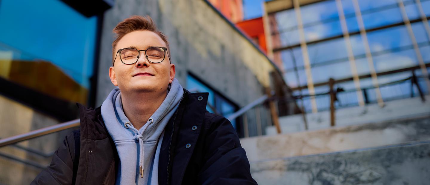 Silmälasipäinen nuori mies hymyilee. Hänellä on silmät kiinni.