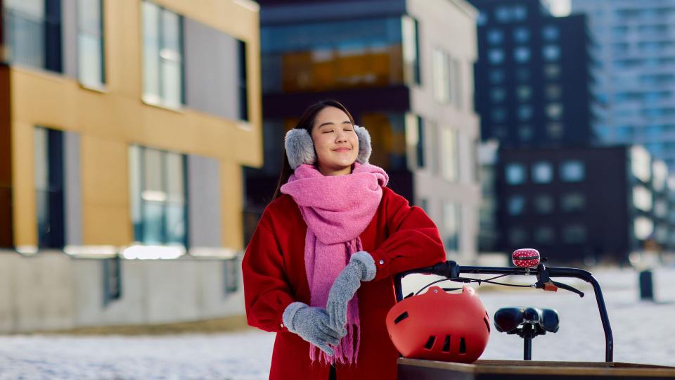Nainen seisoo ulkona talvisäässä punaiseen takkiin pukeutuneena, hymyillen auringonpaisteessa. Hänellä on vaaleanpunainen huivi ja korvalaput, ja edessä on laatikkopyörä.