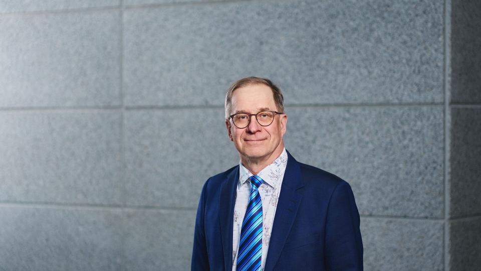 Professori Jouni Pursiainen seisoo hymyillen kiviseinän edessä.