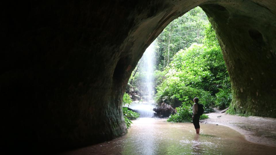 A person standing in an open cave with a background of Amazon waterfall and green forest