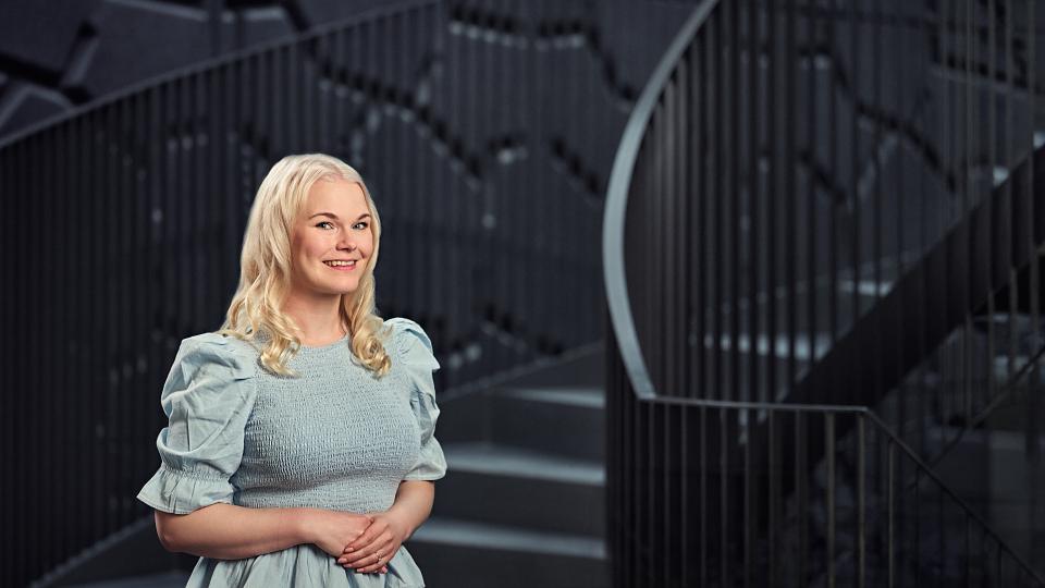 Henkilö, jolla on vaaleat hiukset ja vaaleansininen puffihihoilla varustettu mekko, seisoo sisätiloissa modernin tumman kierreportaan edessä. Taustalla näkyy geometrisia ja nykyaikaisia yksityiskohtia.