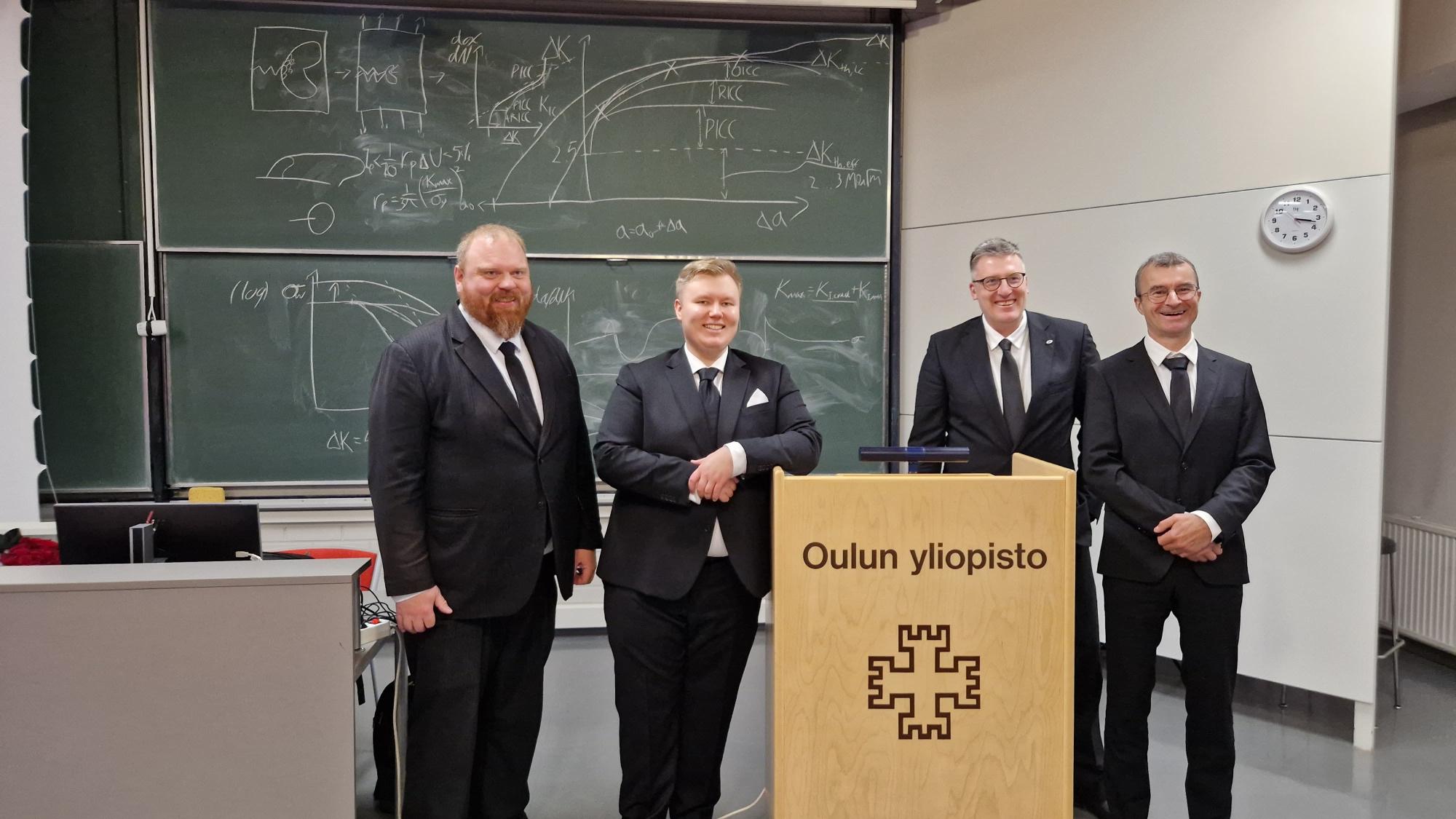 Neljä miestä seisoo hymyillen puhujapöntön takana opetustilassa.