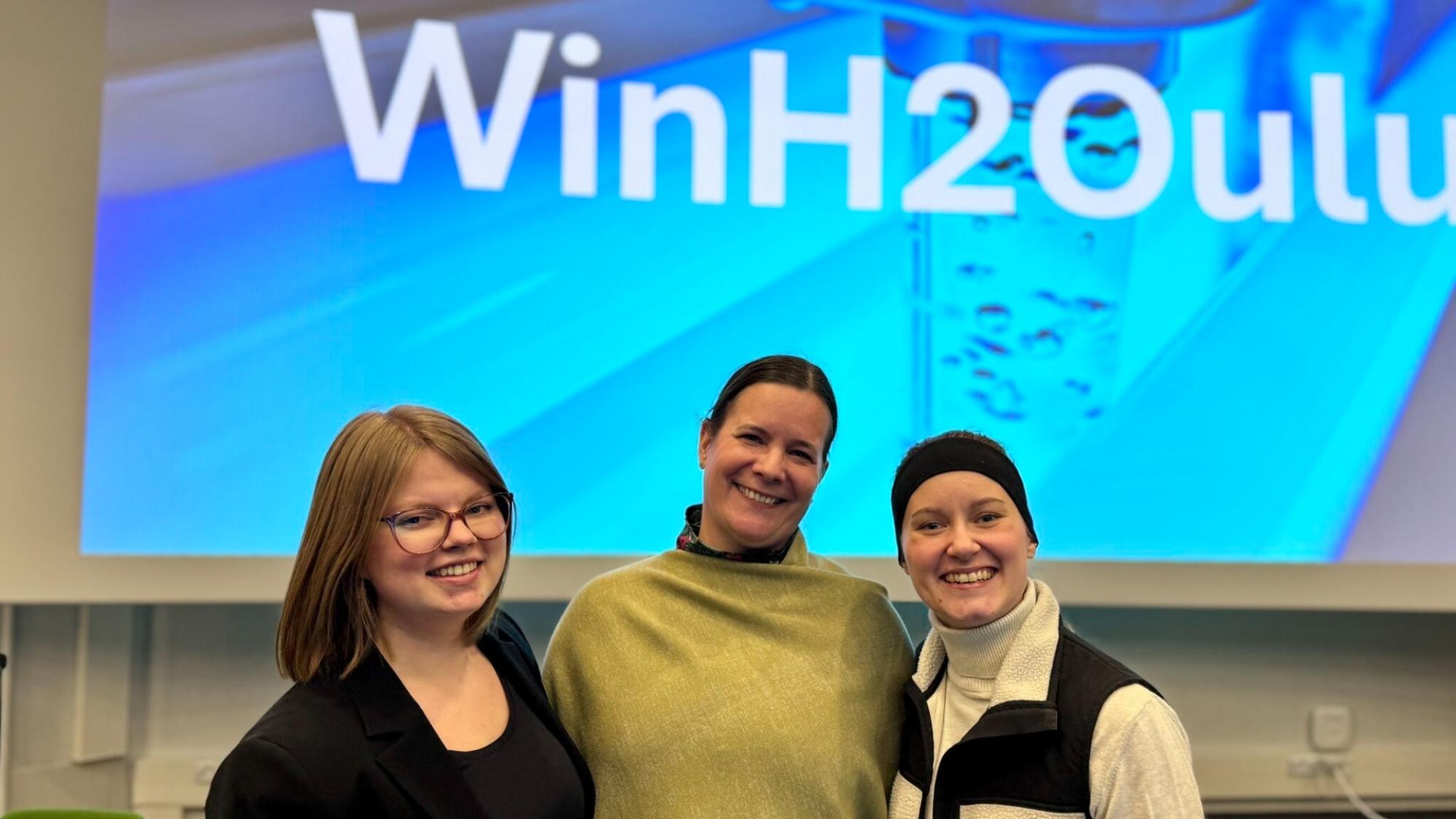 Women smiling in a lecture hall in semi close up photo, screen on the background that reads WinH2Oulu