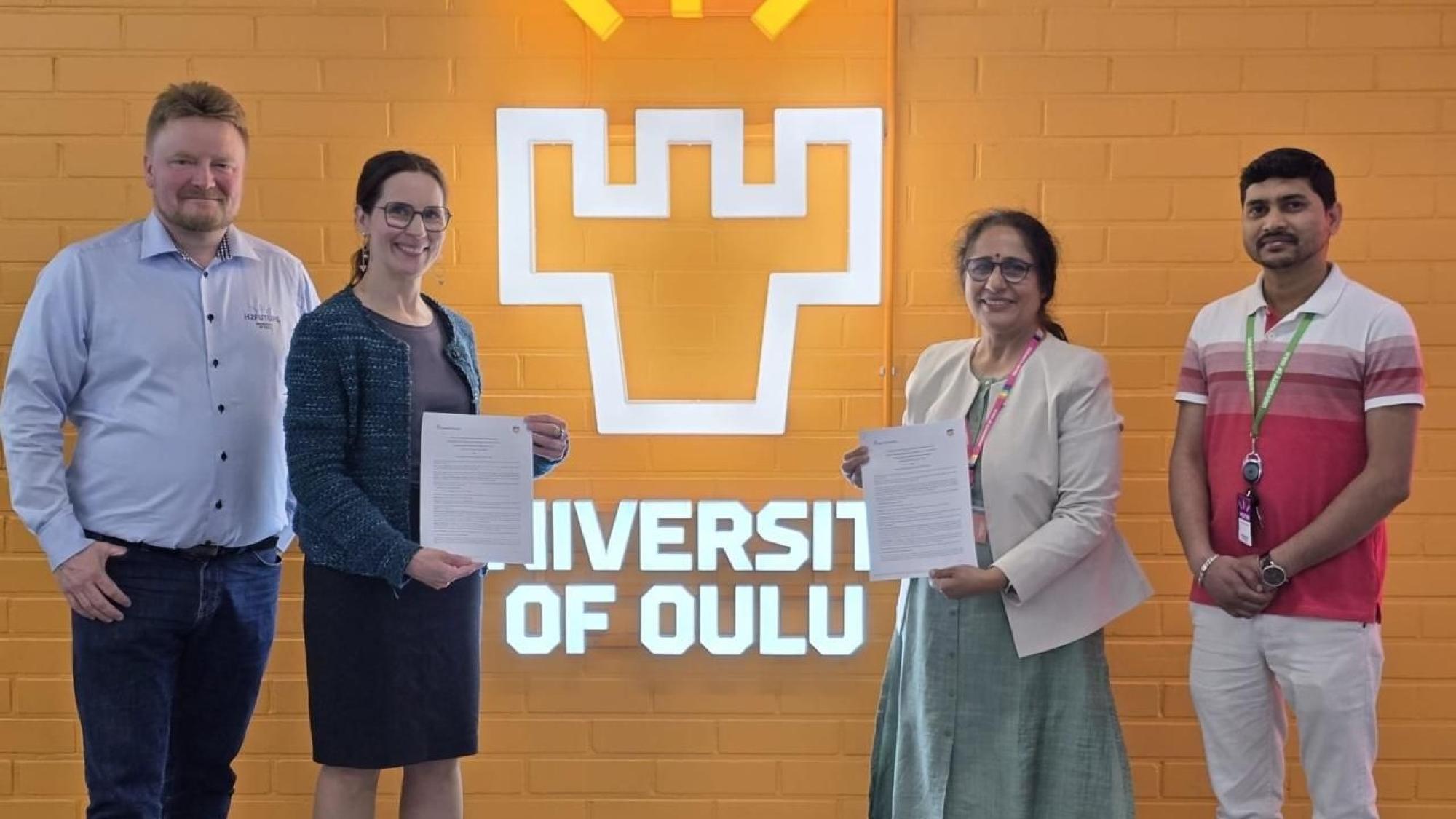 Four persons standing and smiling in front of University of Oulu logo wall
