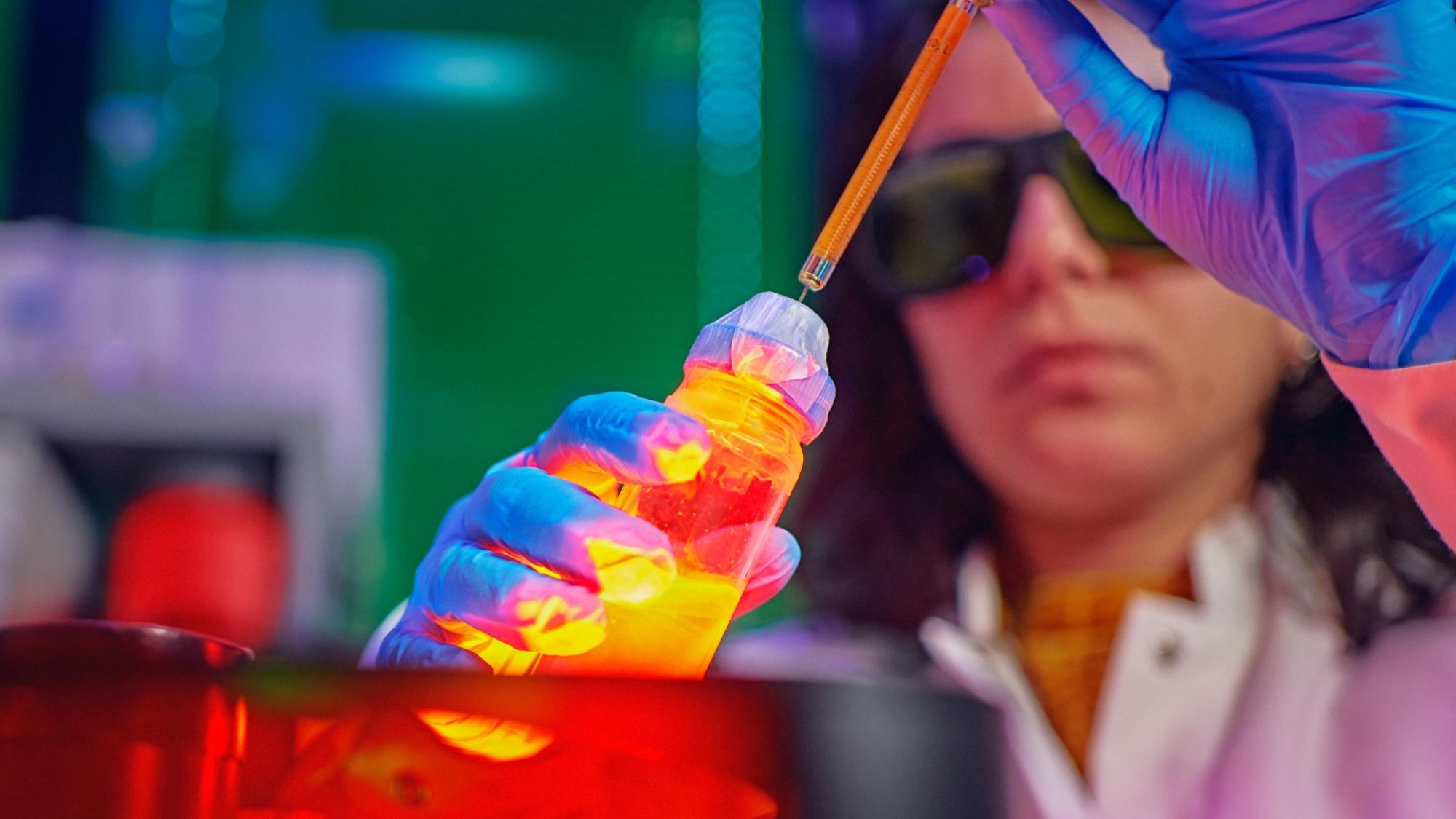 A woman scientist holding a tube with glowing yellow substance in a laboraty with colorful lights