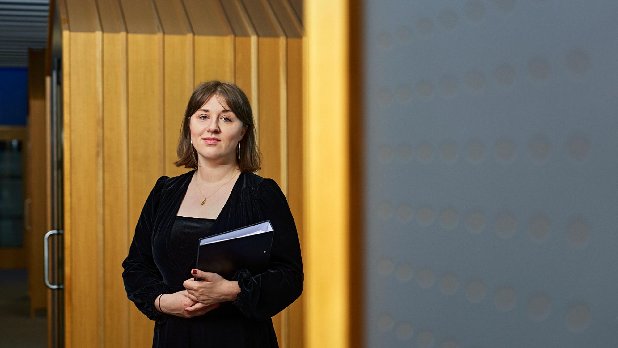 Nainen polkkatukassa seisoo yliopiston käytävällä väitöskirjaa pidellen.