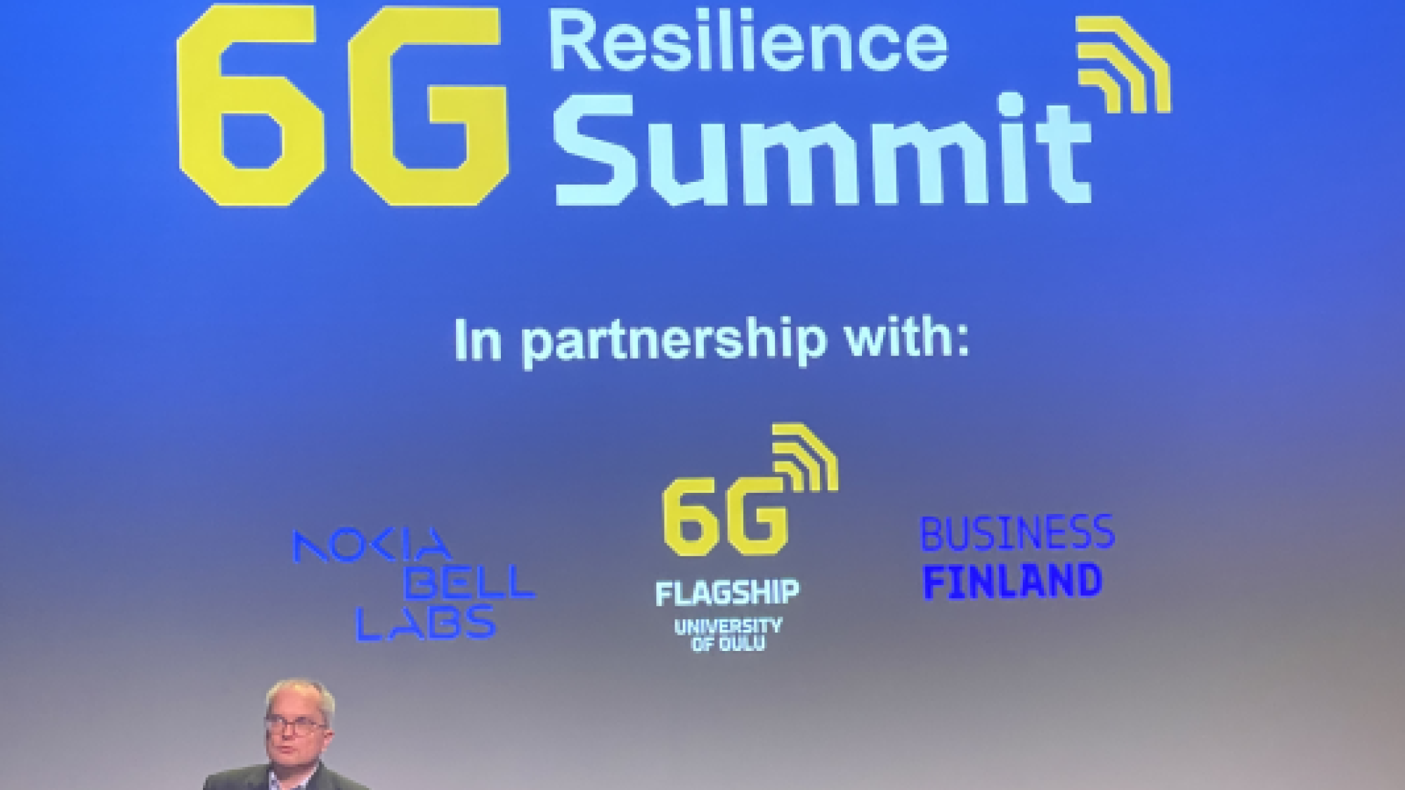 A man giving a speach in front of a display screen. On the screen is a text "6G Resilience Summit".