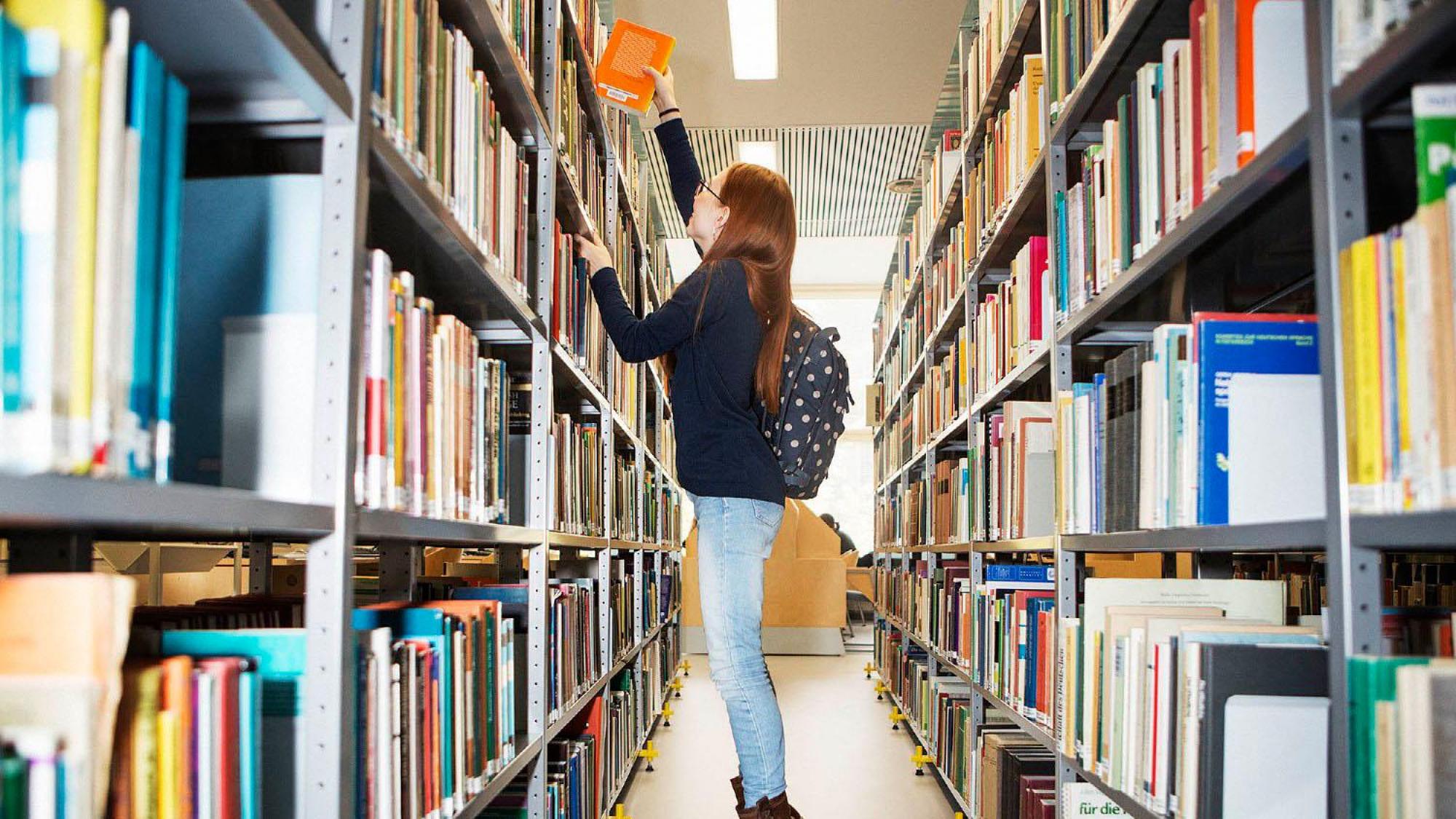 Informaatiotutkimuksen opiskelija seisoo kirjahyllyjen välissä ja ottaa kirjan ylähyllyltä.