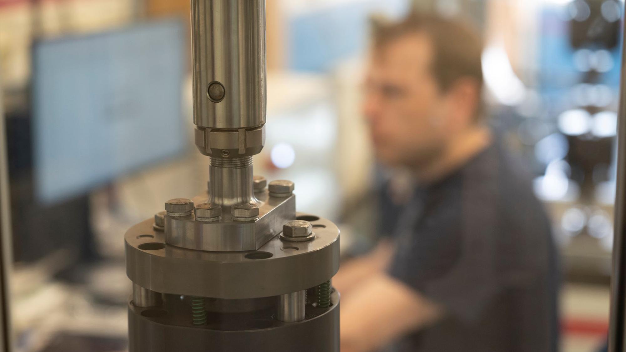 metallin tutkimiseen tarkoitettu laite etualalla, taustalla mies tietokoneen ääressä