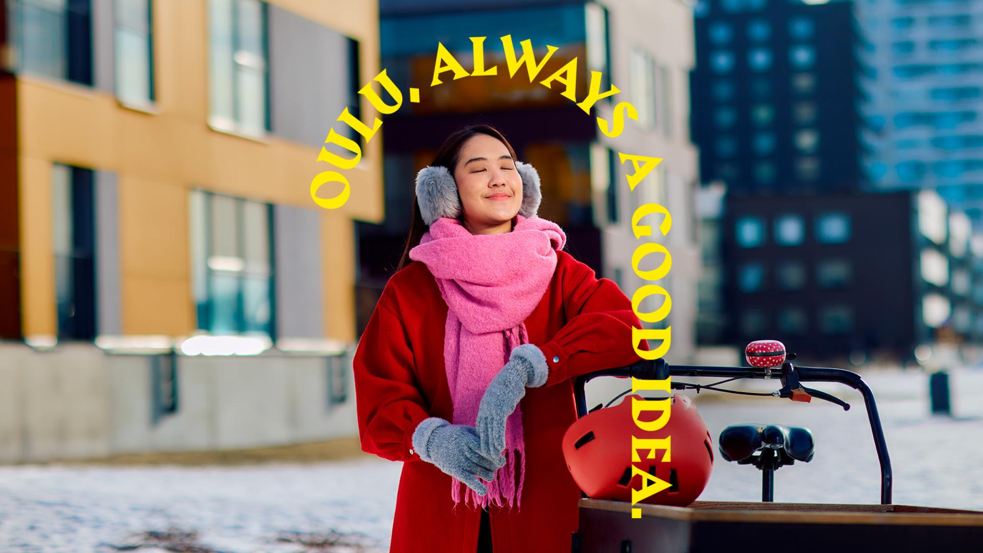 A woman dressed in winter clothing is leaning to a bike trailer. Her eyes are closed. Behind her is a city view. An arrow appears above her stating Oulu, Always a Good Idea.