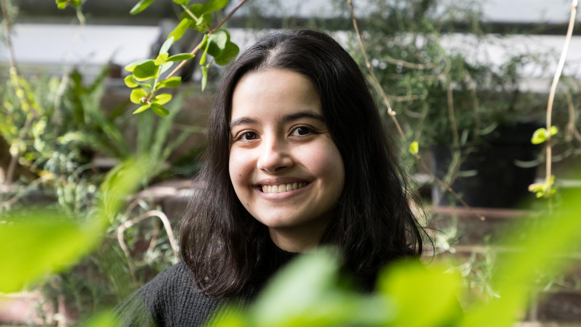 A dark-haired woman is smiling. She is surrounded with green plants.