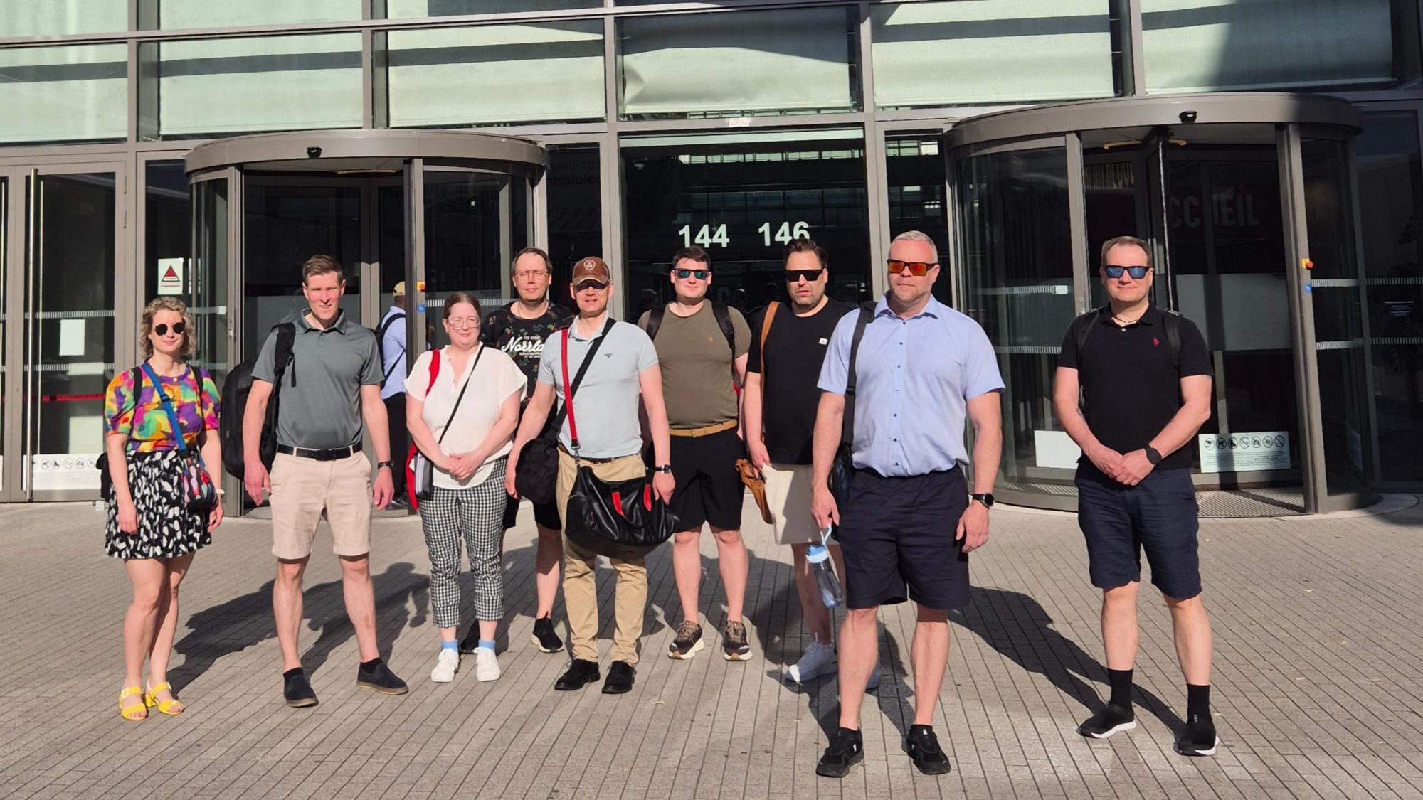 A group of nine people standing in front of a entrance to a building, wearing summer clothes.