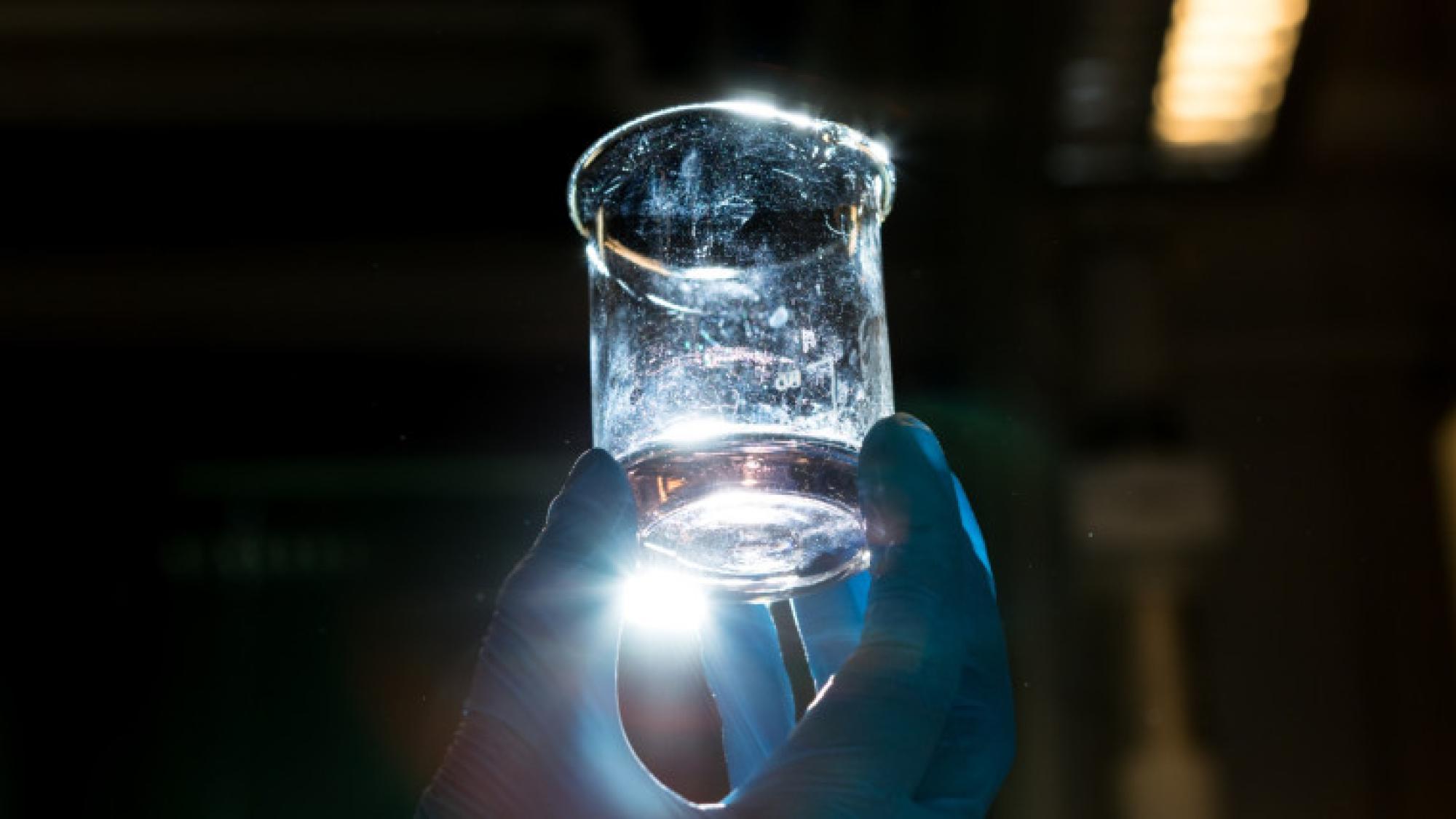 A hand with protective gloves holding a measuring cup with fluid up against light
