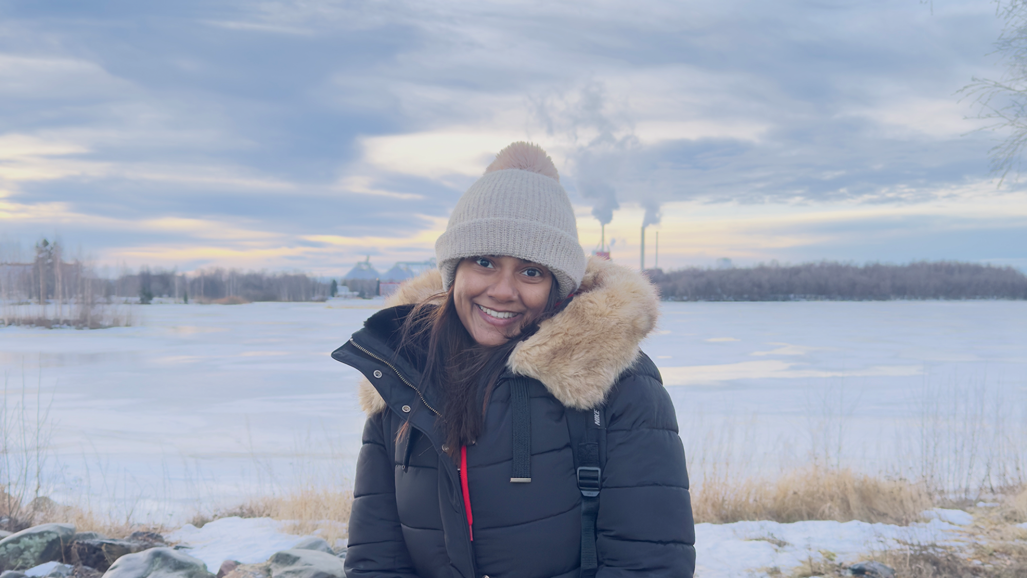 A young woman smiling while posing in a snowy seaside with a landscape on the opposite shore.