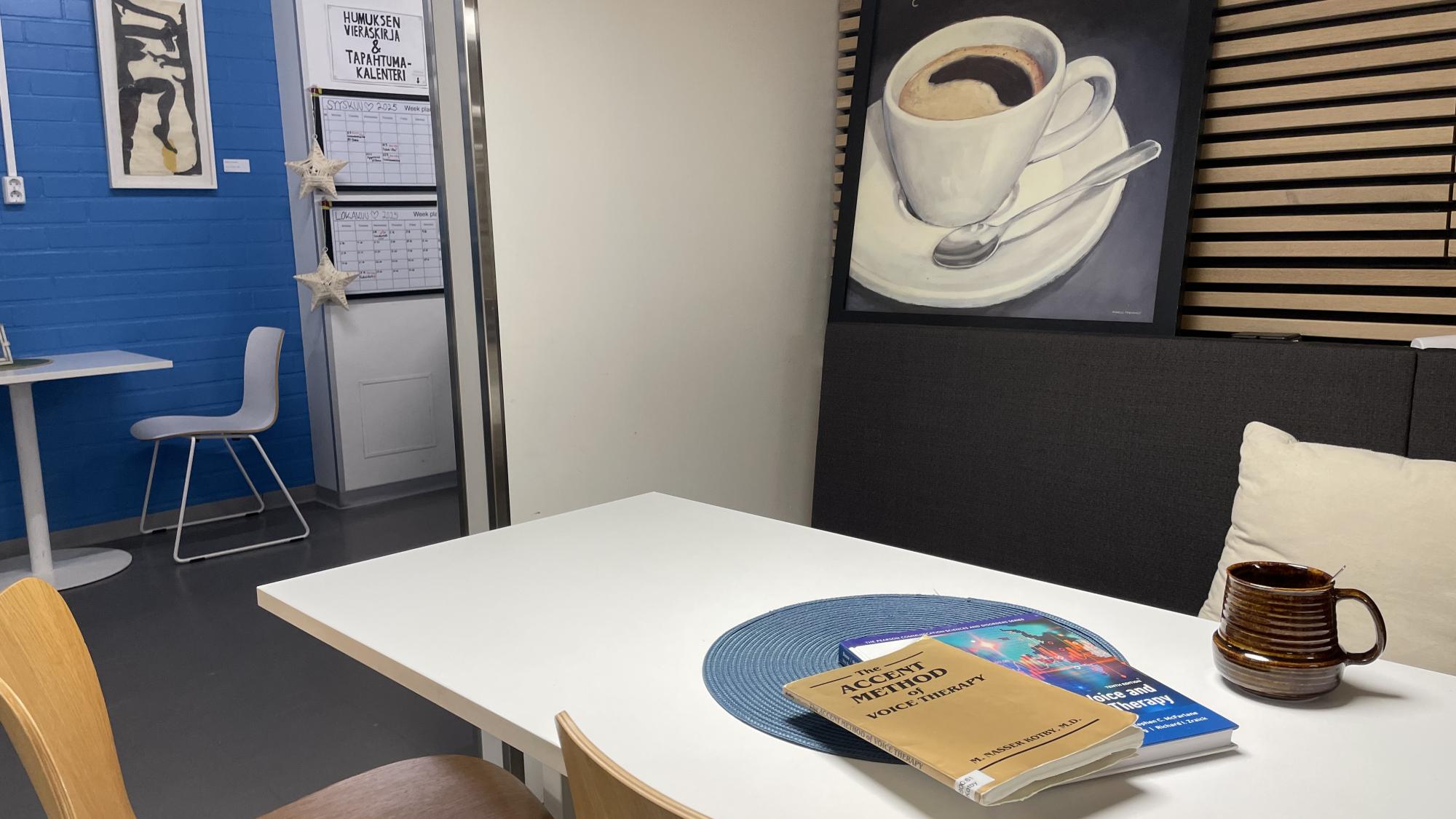 Kuva Oulun yliopiston Humus-kuppilasta. Kuvassa on pöytä ja tuoleja, ja pöydän päällä on kirjoja sekä teekuppi. Seinällä on kuva kahvikupista.