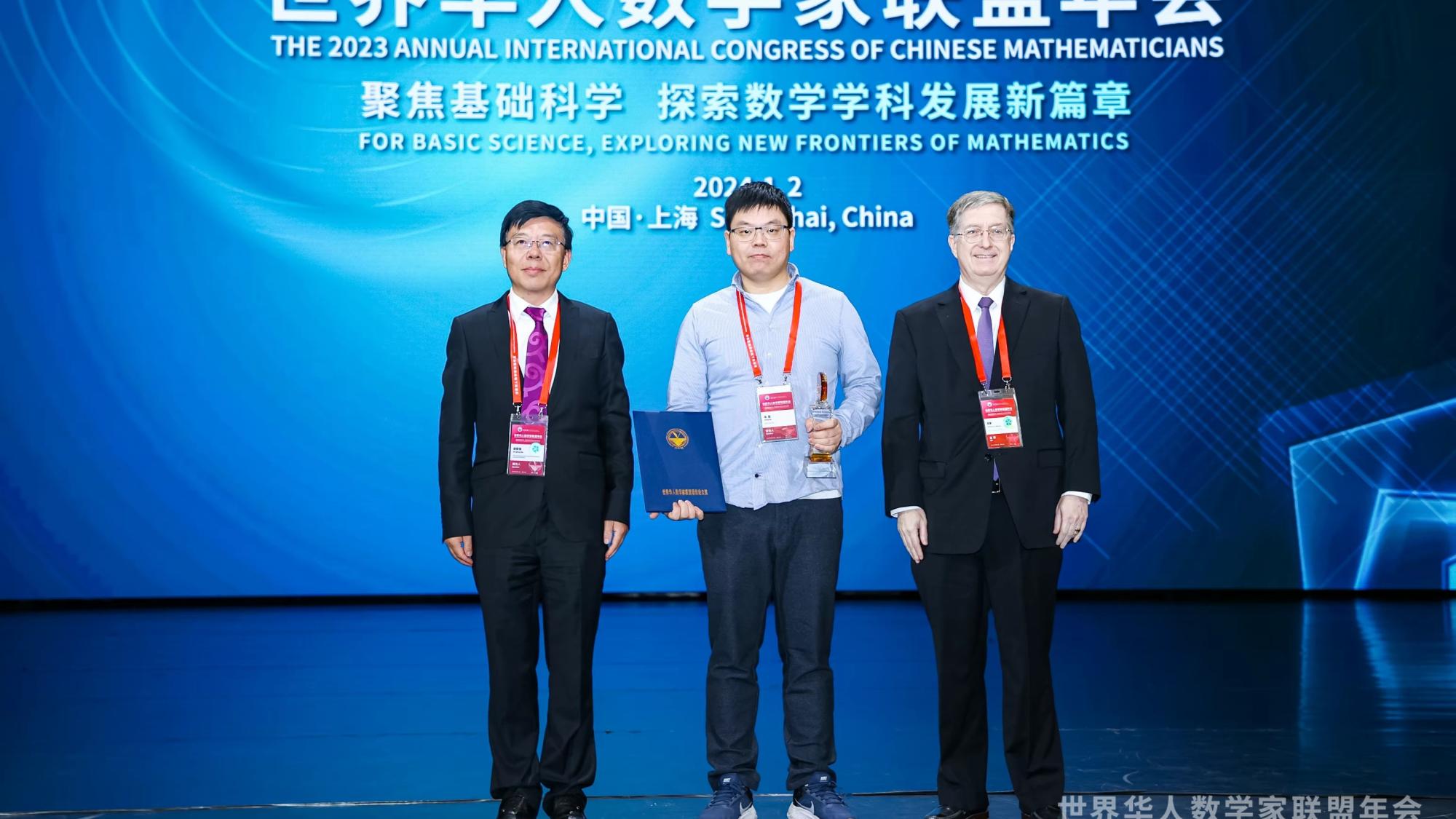 Meng Wu, an Associate Professor of Mathematics at the University of Oulu, holds the 2023 International Consortium of Chinese Mathematicians Best Paper Award at the center of a stage in Shanghai, China.