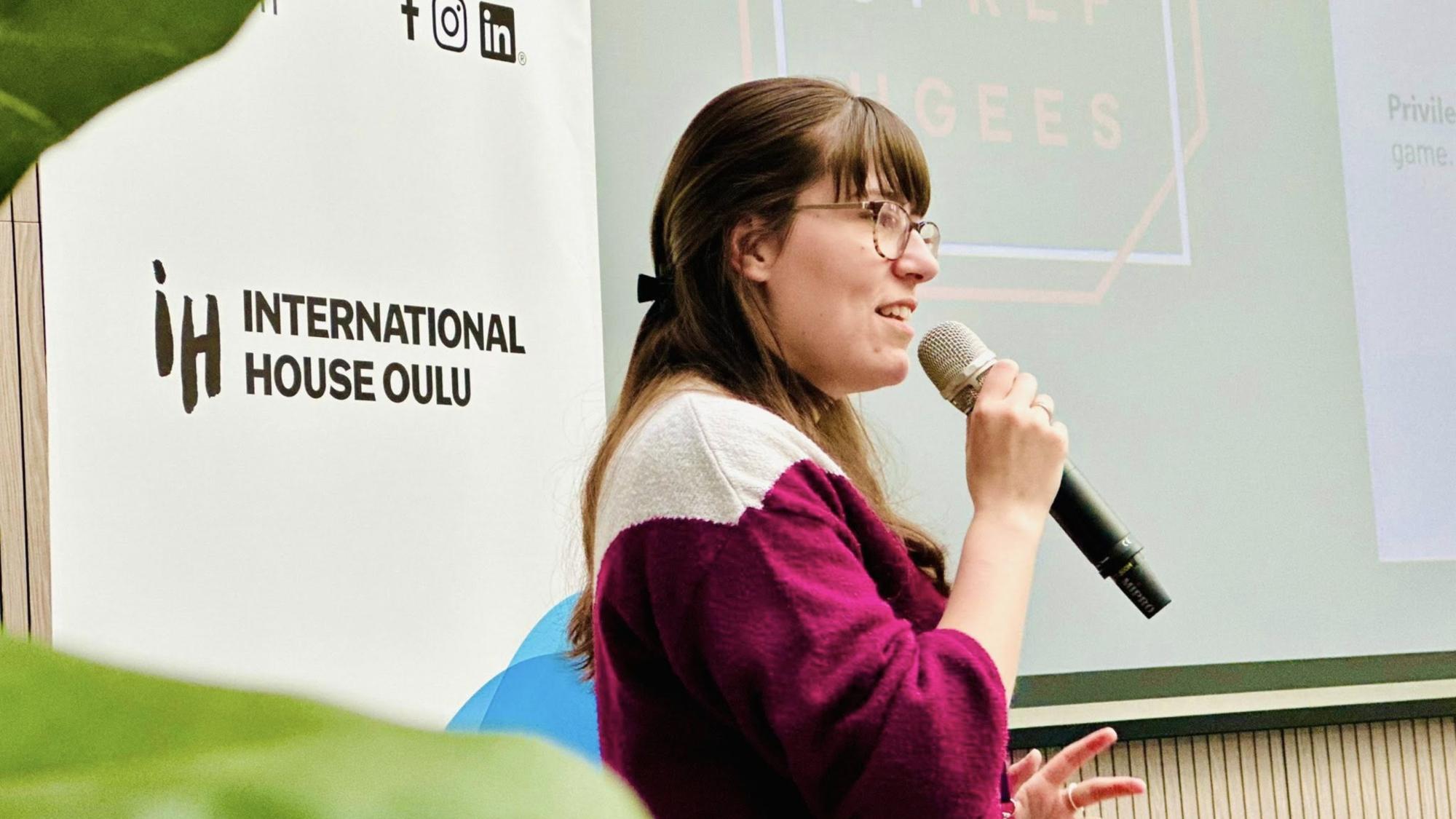 A girl with dark hair and glasses holding a microphone to her mouth in front of a banner that says ‘International House Oulu’.