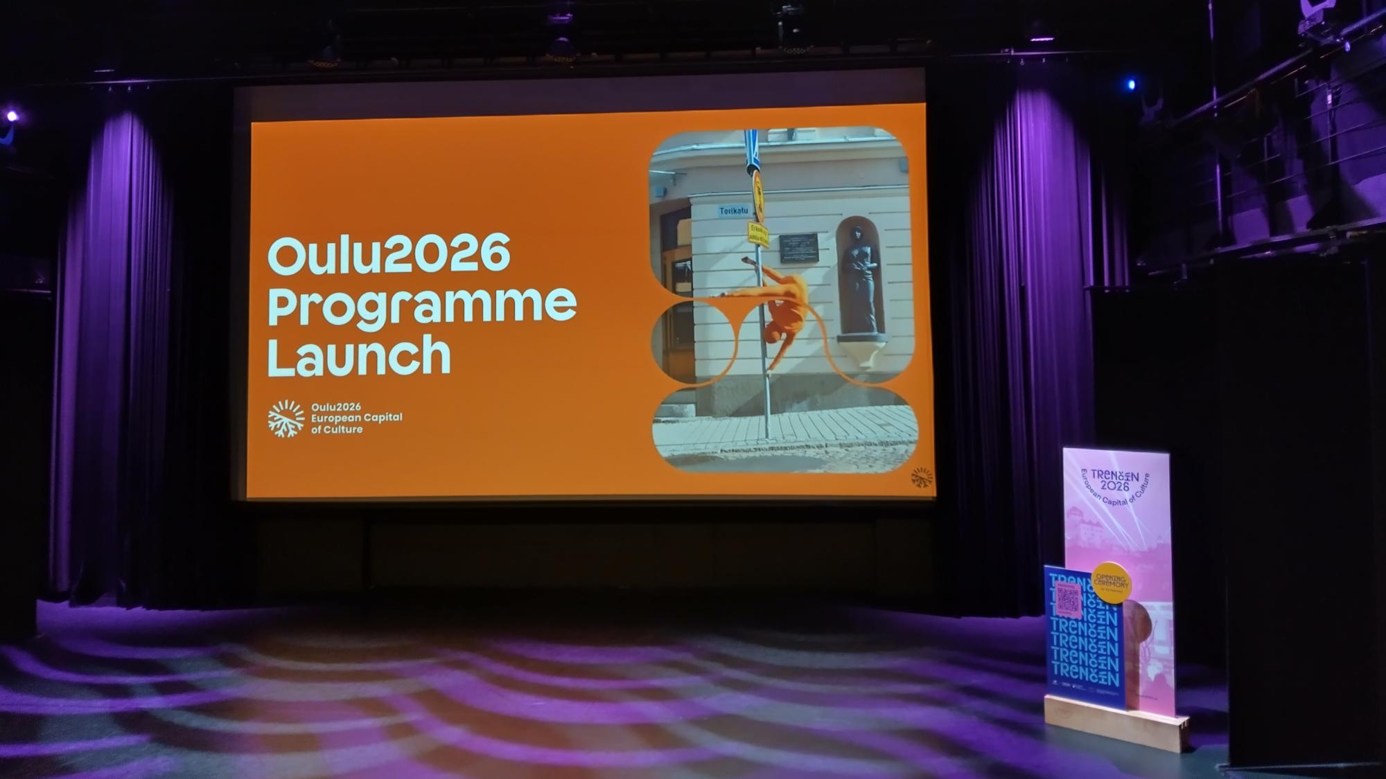 Oulu2026 julkistustilaisuuden iso valkokangas Valvesalissa, violetit tunnelmavalot taustalla.