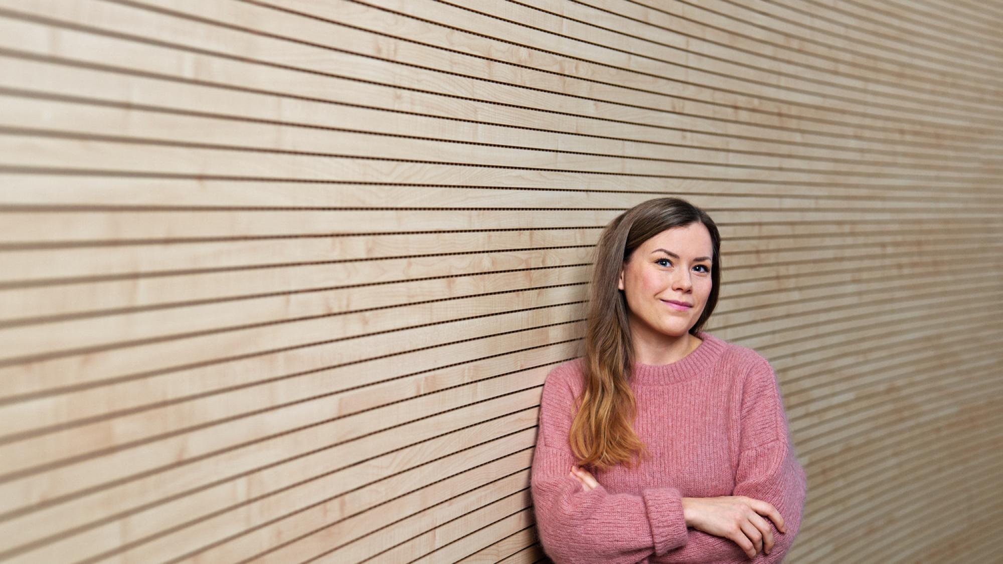 Arla Magga in portrait photo, a light pink sweater on and wooden wall as a background.