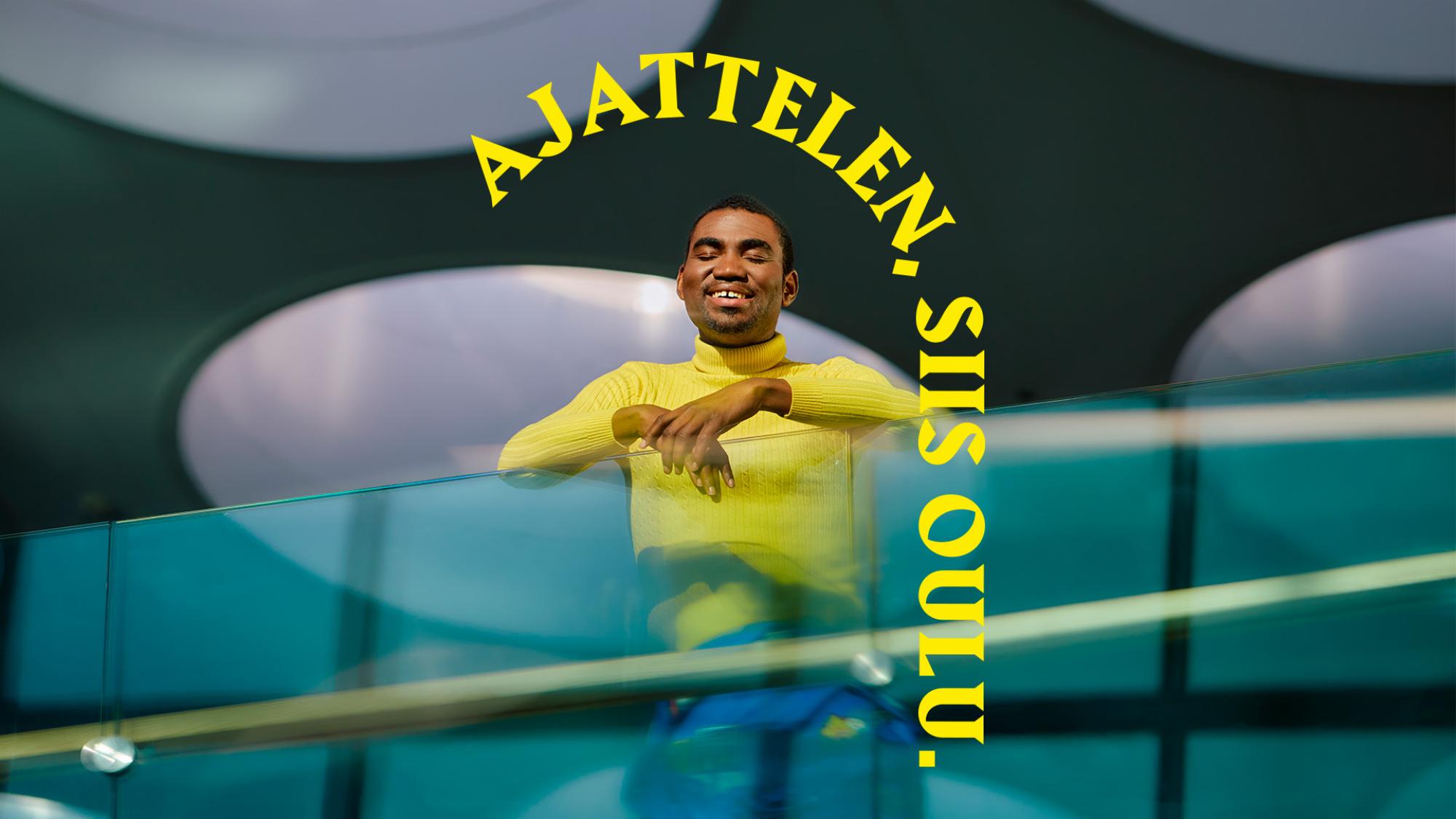 Keltapaitainen mies nojaa silmät kiinni lasikaiteeseen. Hänen takanaan on pyöreitä kattoikkunoita. Päähenkilön yläpuolella on keltainen kaari, jossa lukee Ajattelen, siis Oulu.