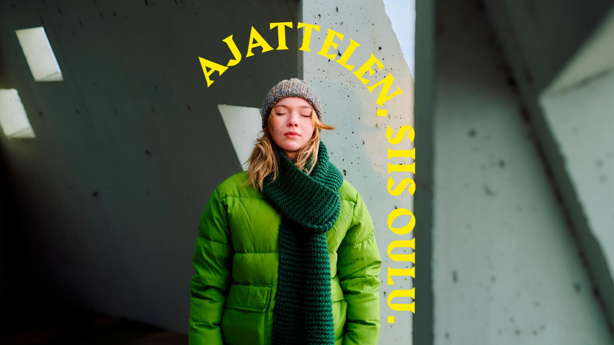 Huivi- ja pipopäinen nainen vihreässä takissa seisoo silmät kiinni pylvään edessä. Hänen takanaan on betoniseinää. Päähenkilön yläpuolella on keltainen kaari, jossa lukee Ajattelen, siis Oulu.