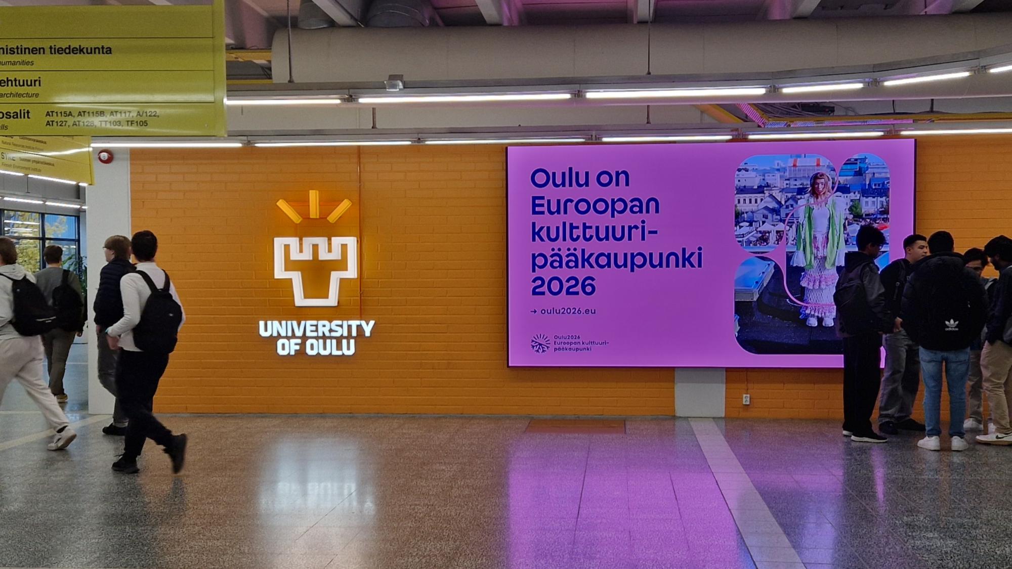 An image of people walking on campus with an advertisement of Oulu capital of culture displayed in the background