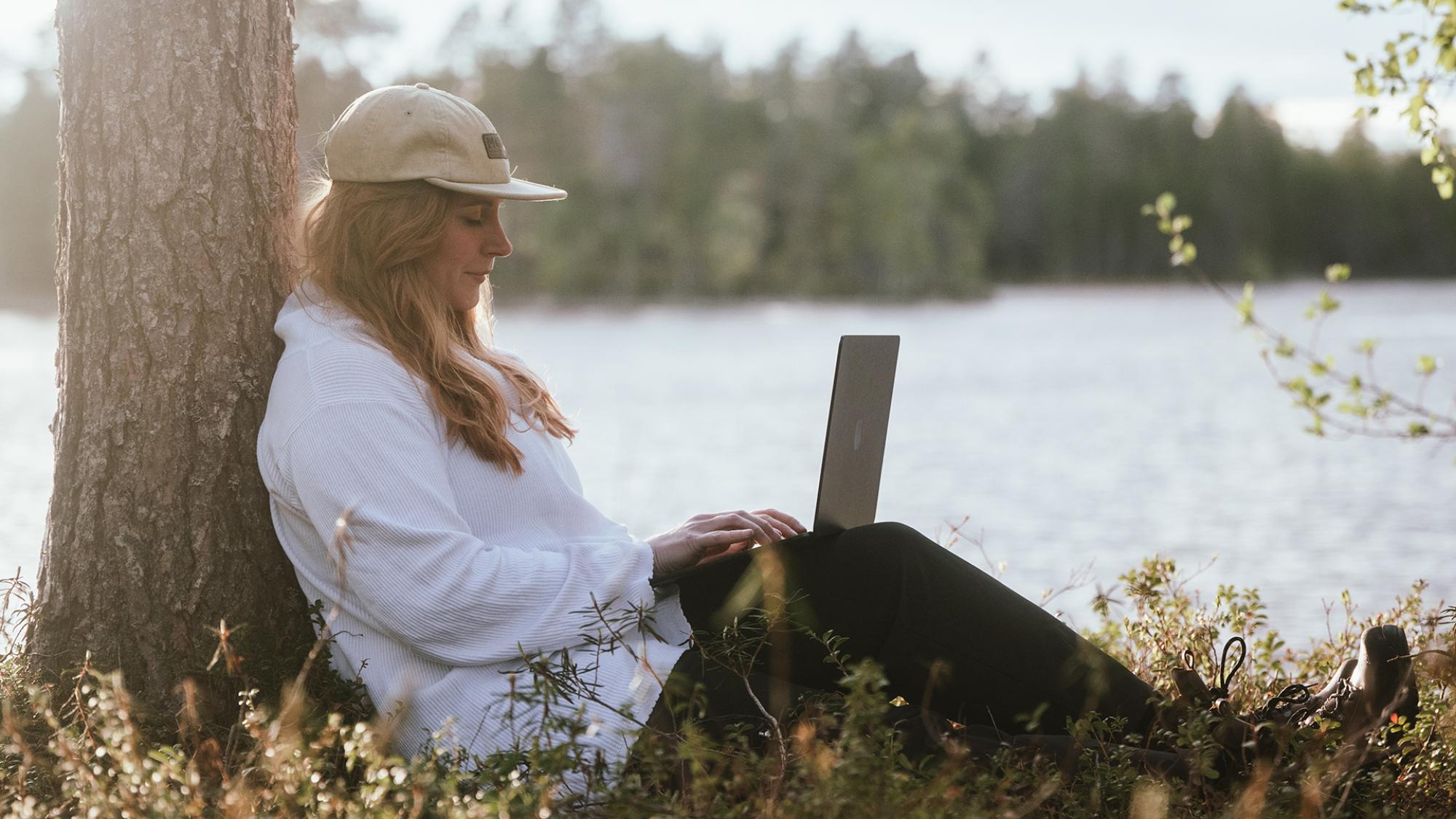 nainen istuu puun juurella järven rannalla läppäri sylissä