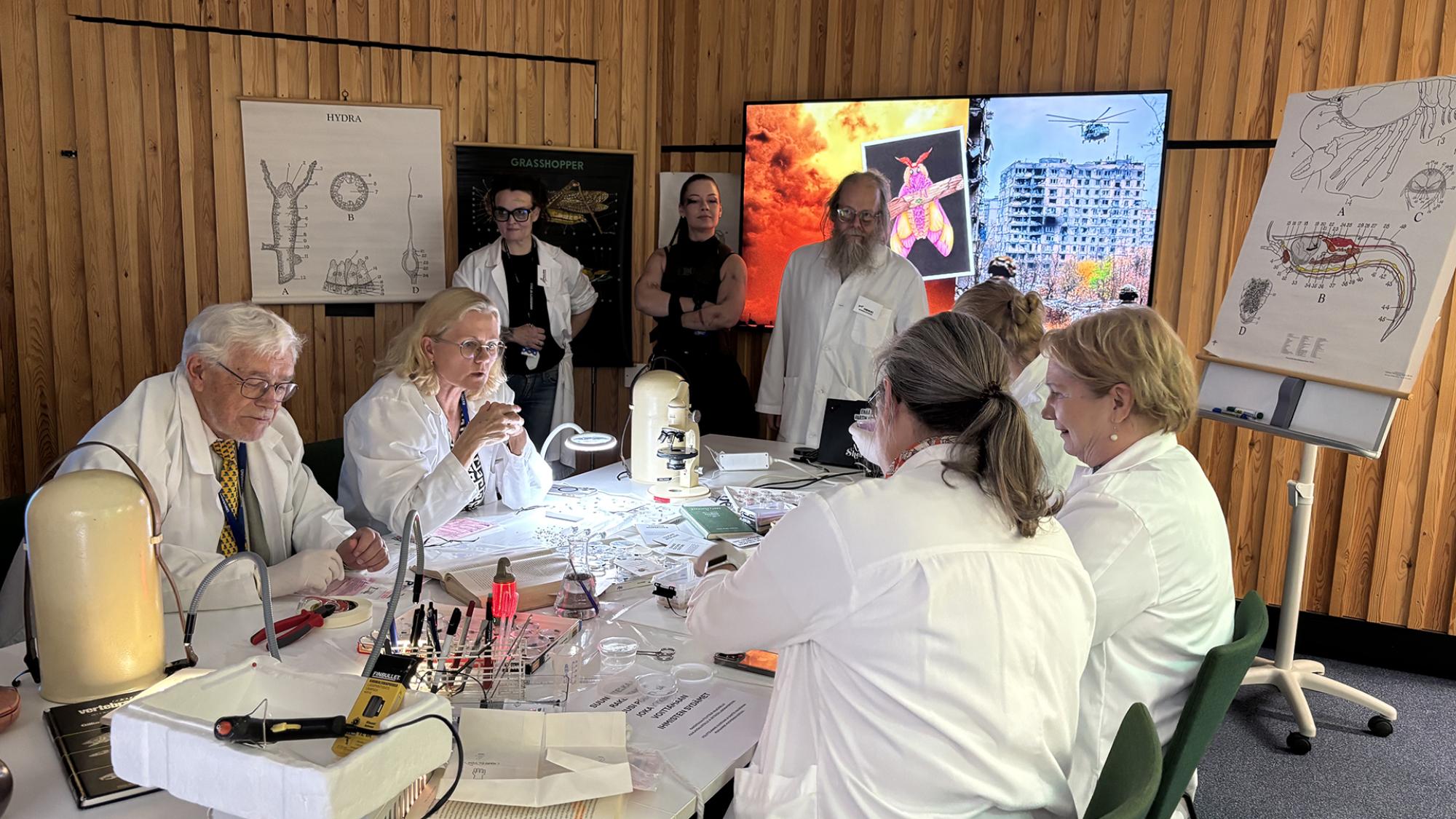 Ihmisiä valkoisissa takeissa työskentelemässä pöydän ääressä. Pöydällä on paljon erilaista työskentely- ja tutkimusvälineistöä, mm. mikroskooppeja, petrimaljoja ja kirjoja.