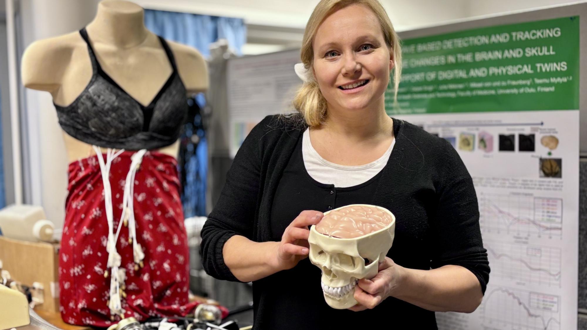 Senior researcher Mariella Särestöniemi in the Human Body Twin Laboratory, where technologies for portable and wearable health monitoring are developed and evaluated. She holds a head phantom used to study signal propagation in brain tissue, while a wearable breast cancer detection vest is displayed on a mannequin in the background.
