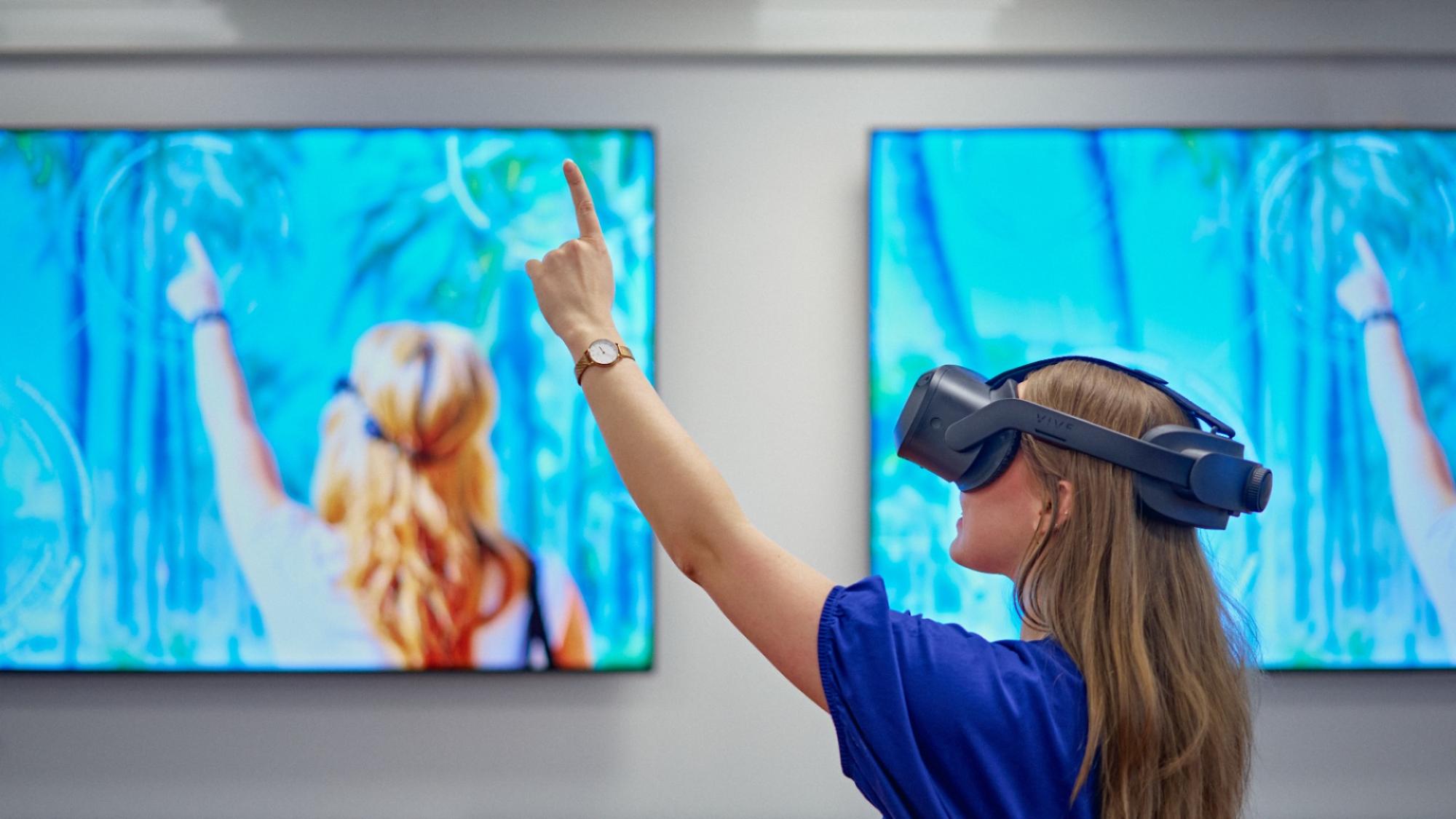 A young women with VR goggles facing a blue screen and pointing at it