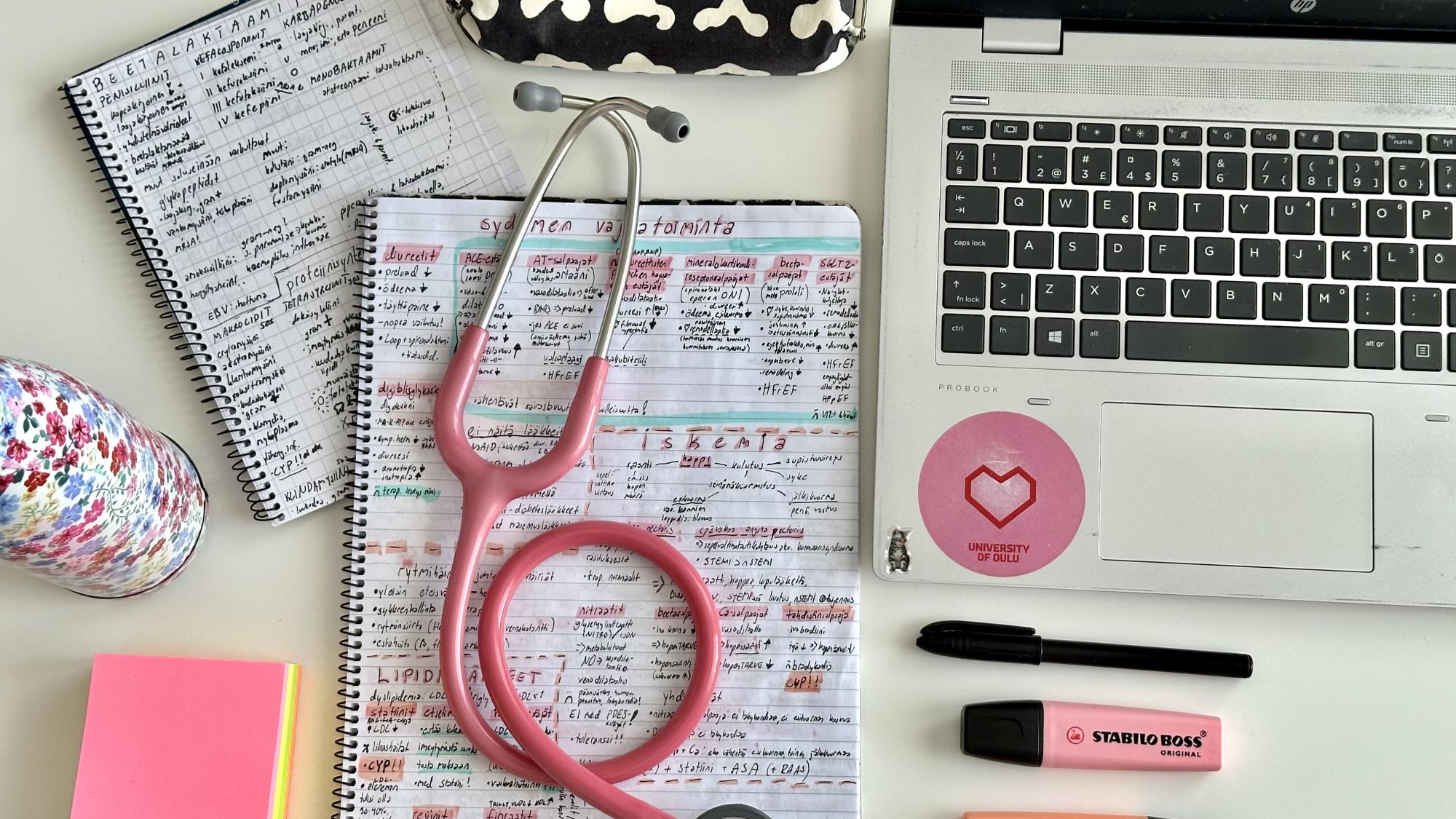 Kuvassa on tietokone, punertavia yliviivaustusseja, värikkäitä muistiinpanolappuja, pilvikuvioitu penaali, kukkakuvioitu juomapullo ja kaksi muistiinpanovihkoa, joista isomman päällä on pinkki stetoskooppi.