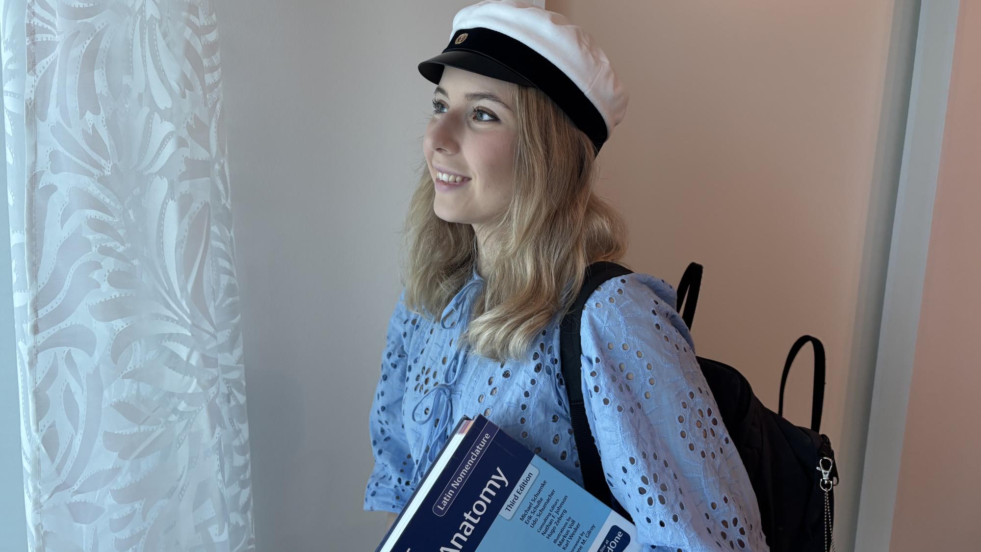 Nuori nainen katsoo ikkunasta ulos. Hänellä on ylioppilaslakki päässä, kirja kädessä ja reppu selässä.