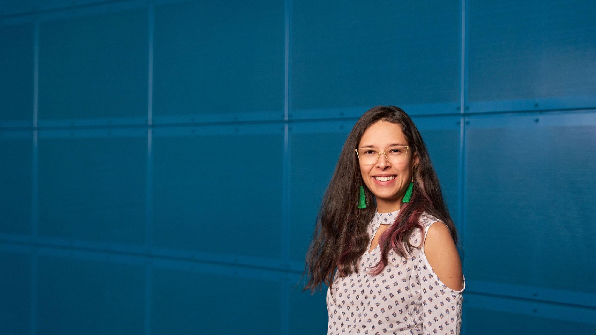 A person with mid long brown hair looks to te camera and smiles. Blue background. 
