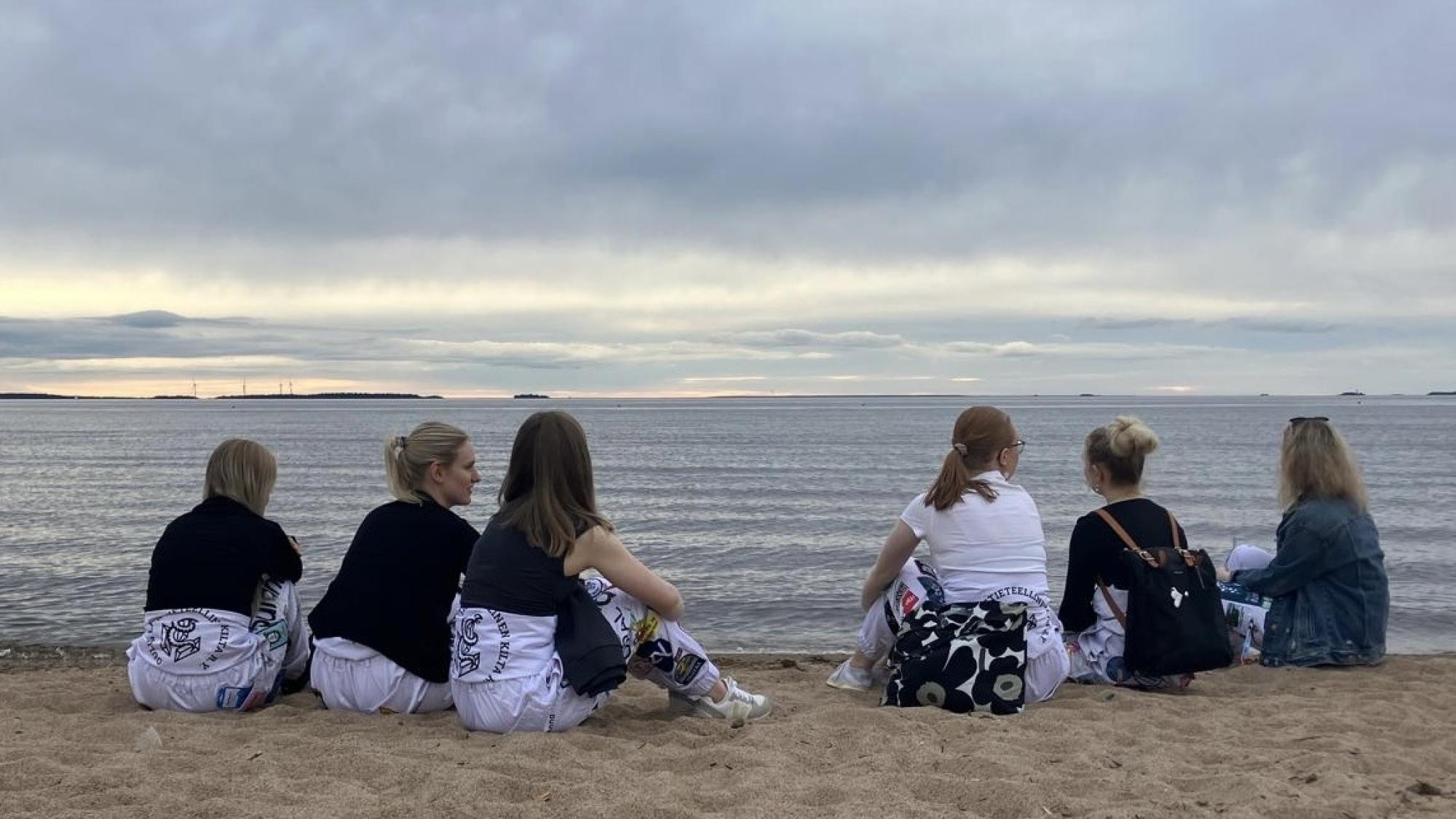 Kuusi opiskelijaa istuu hiekassa merenrannalla. He ovat selin ja katsovat kohti merinäkymää. Heillä on yllään valkoiset opiskelijahaalarit.