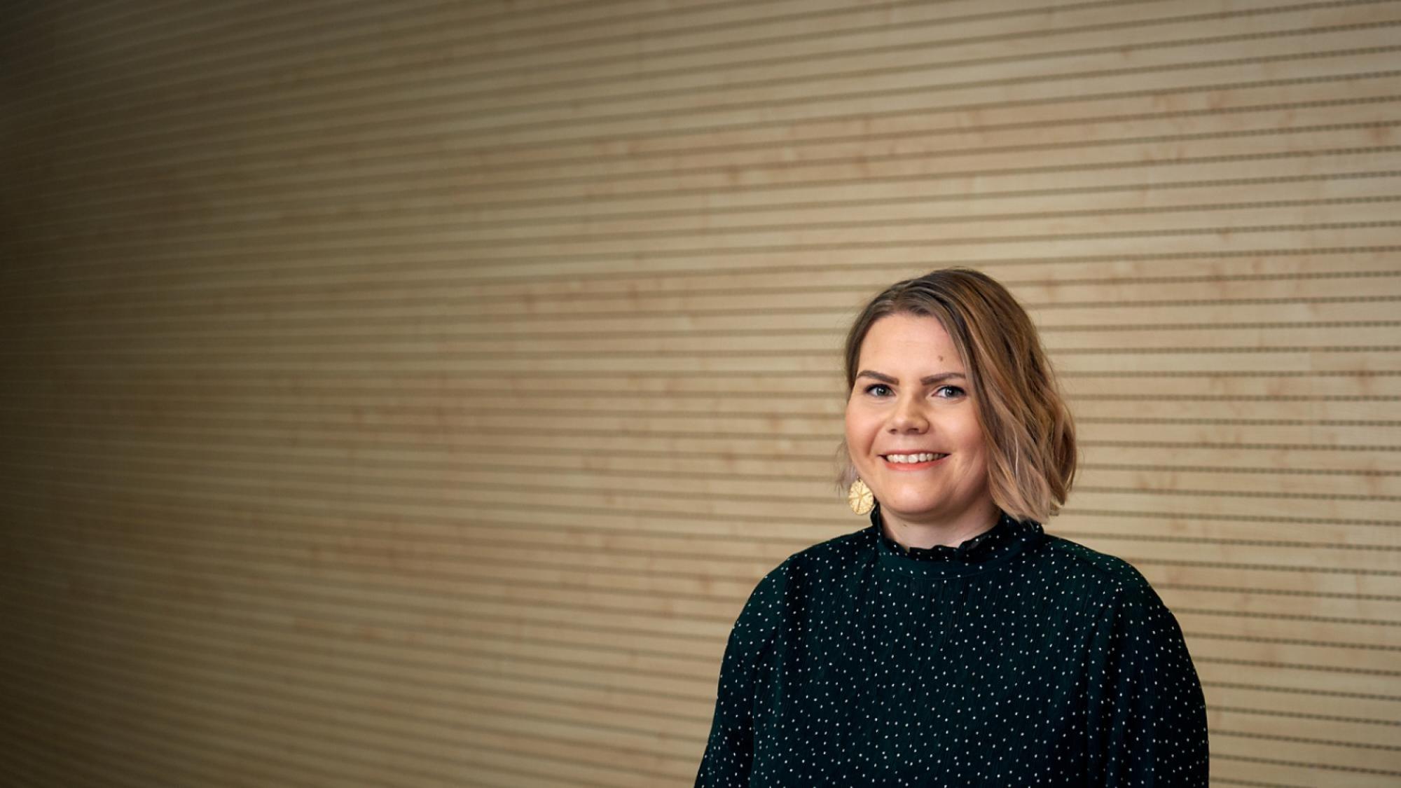 Postdoctoral researcher Tiina Törmänen smiling in portrait photo with light wooden wall in the background