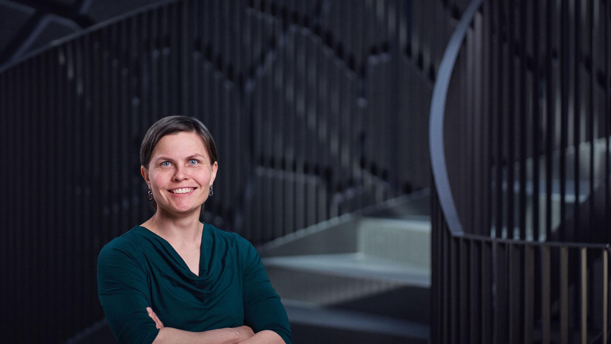 Portrait of Salla Kangas, a dark-haired woman wearing a green shirt, smiling warmly while standing on a stairway. The setting is bright and professional.