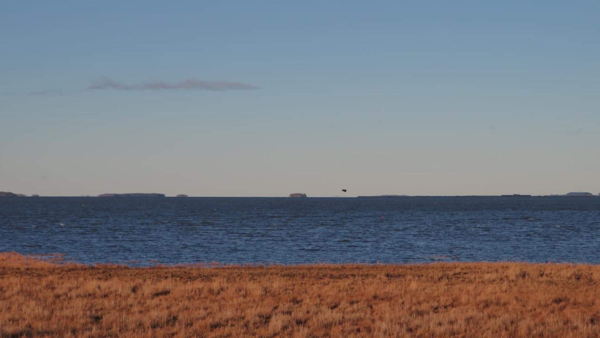 A sea view from a shore. A bird is flying above the sea.