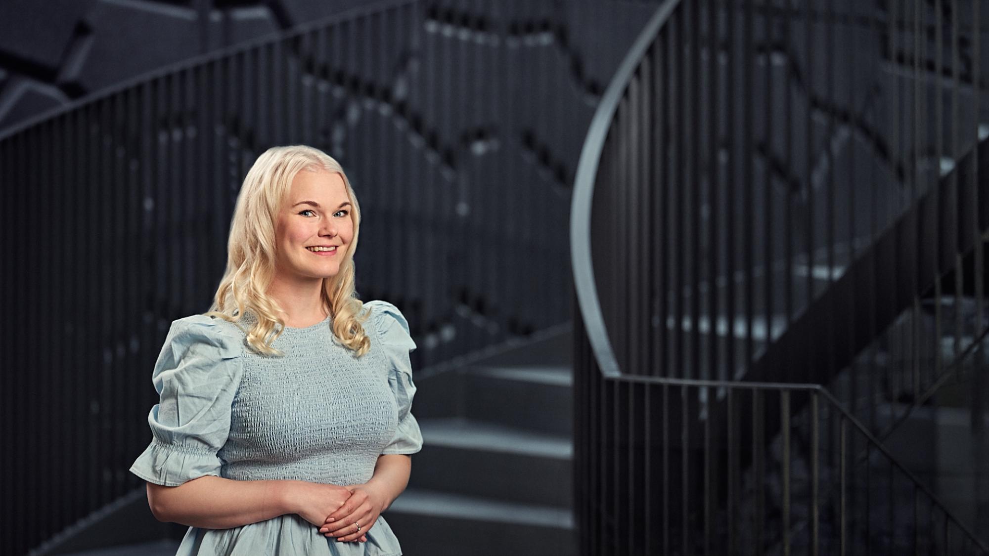 Henkilö, jolla on vaaleat hiukset ja vaaleansininen puffihihoilla varustettu mekko, seisoo sisätiloissa modernin tumman kierreportaan edessä. Taustalla näkyy geometrisia ja nykyaikaisia yksityiskohtia.