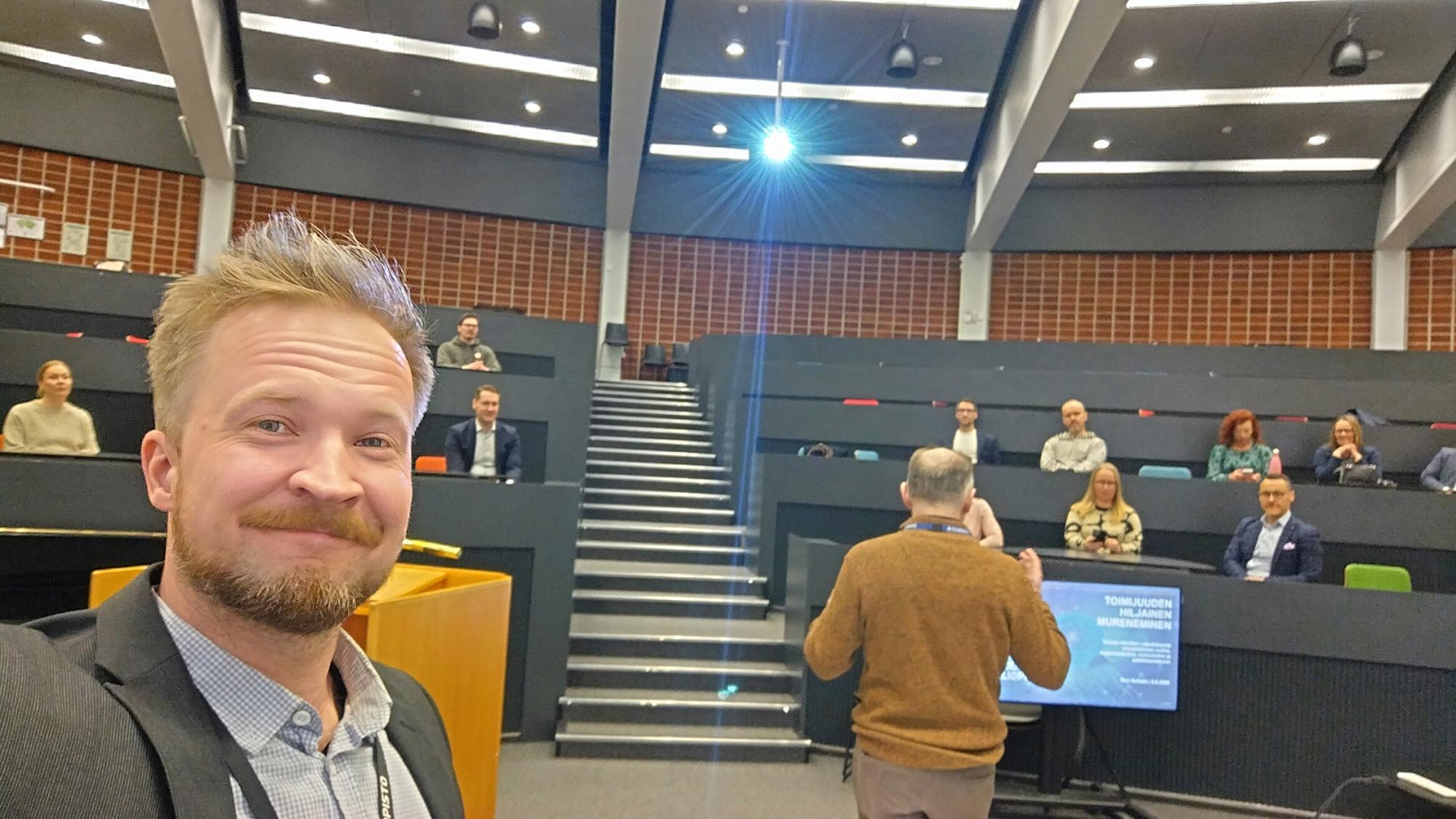 A smiling man looking at the camera in front of an audience.
