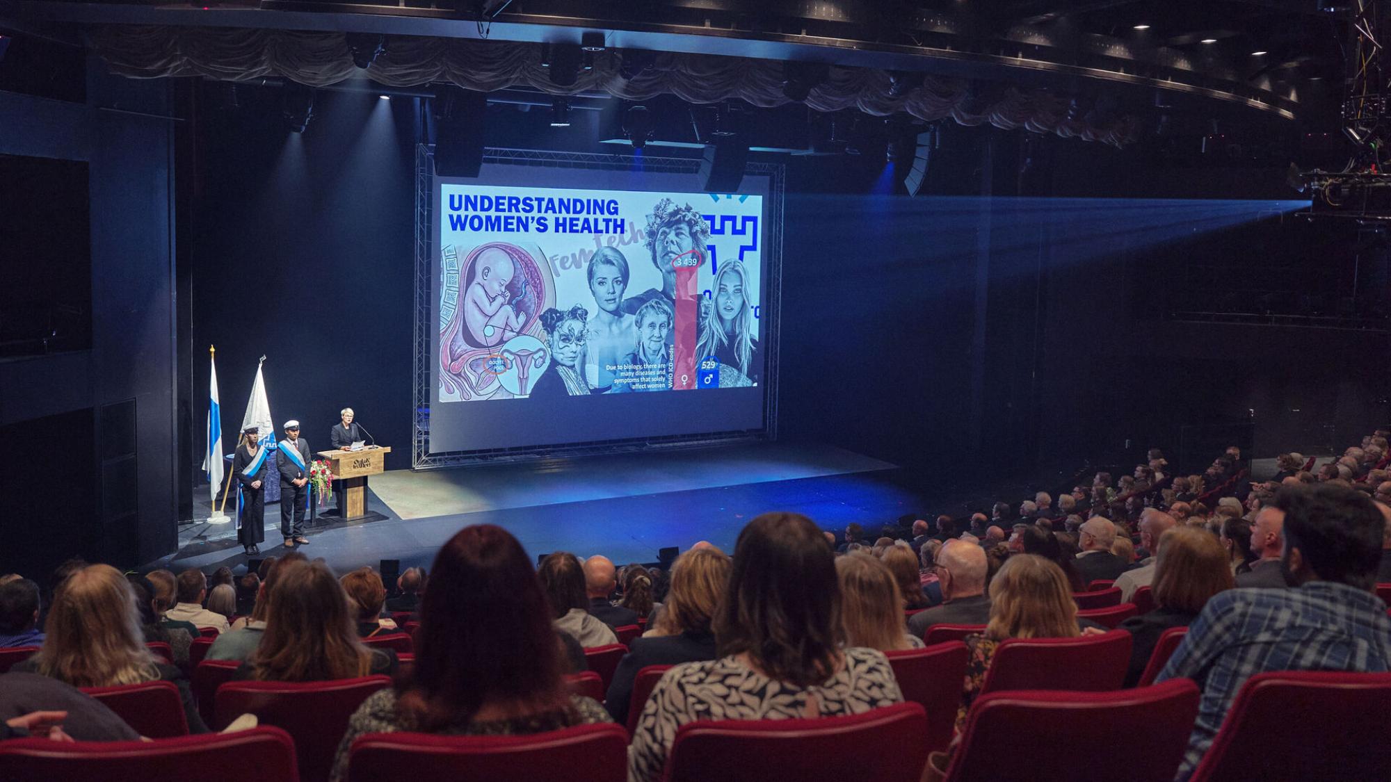 Yleisöä teatterin katsomossa, valkokankaalla esitys naisten terveydestä.