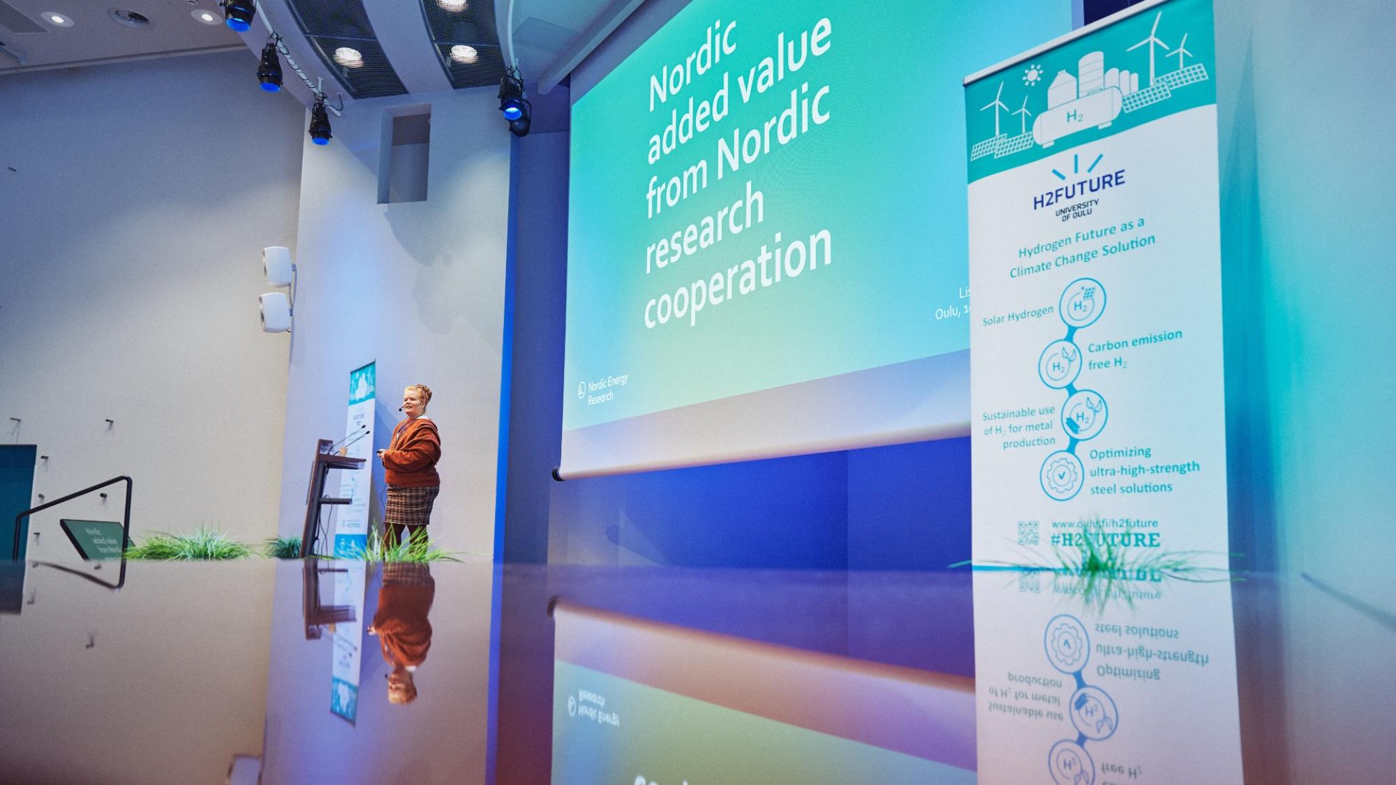 A woman giving a presentation on a big stage with blue and turquose screen on the background and everything reflected like a mirrow on a surface in the below