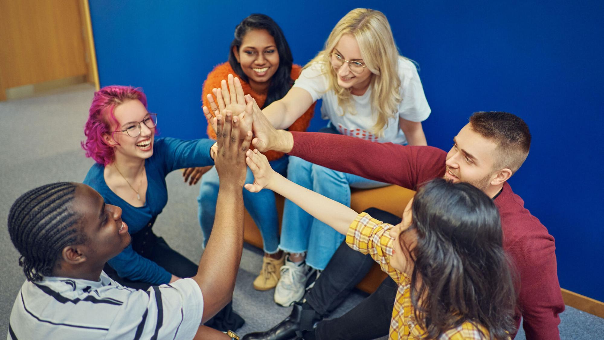 Smiling, multinational student group giving a high five with a blue wall on the background