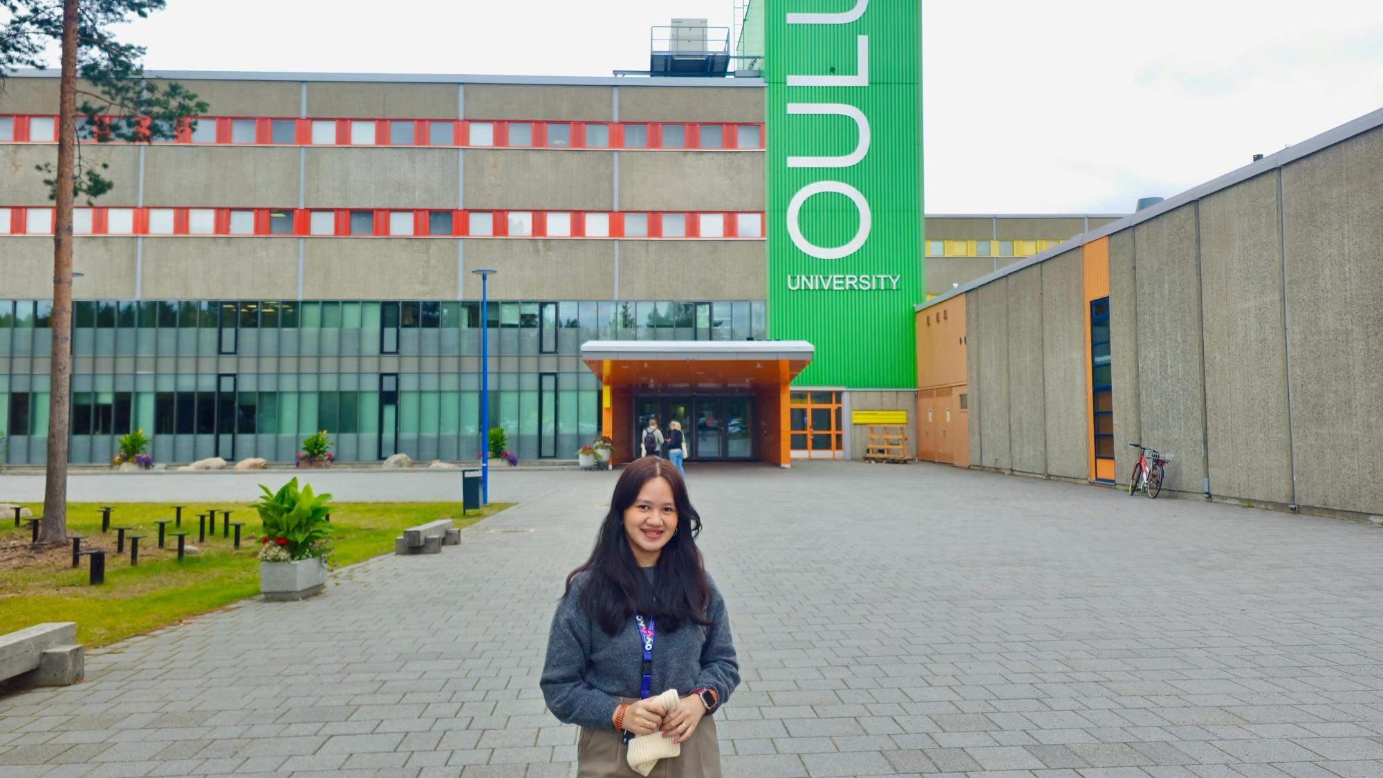 A brown-haired girl is standing in front of a concrete building. A sign stating Oulu University is visible on the green wall.