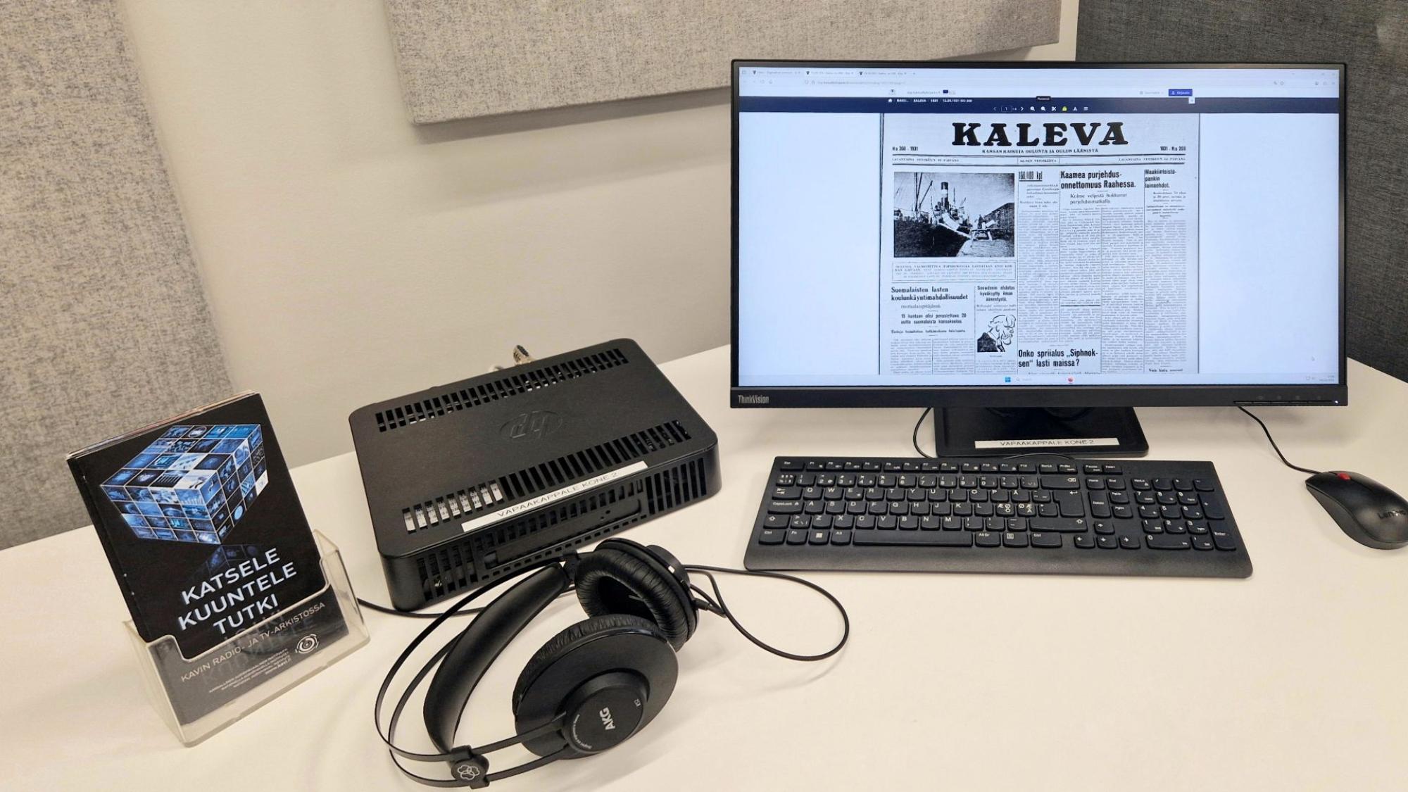 Tiedekirjasto Pegasuksen vapaakappaletyöasema: pöydällä tietokone, näppäimistö, näyttö, hiiri ja kuulokkeet sekä esitteitä. Vapaakappaletyöaseman näytöllä näkyy Kalevan etusivu vuodelta 1931.