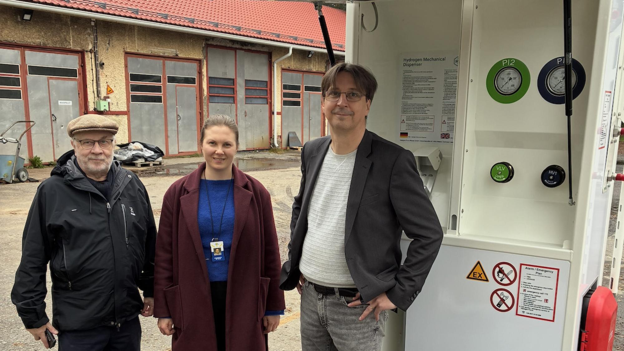 Three people standing in front of a Hydrogen refuelling equipment.