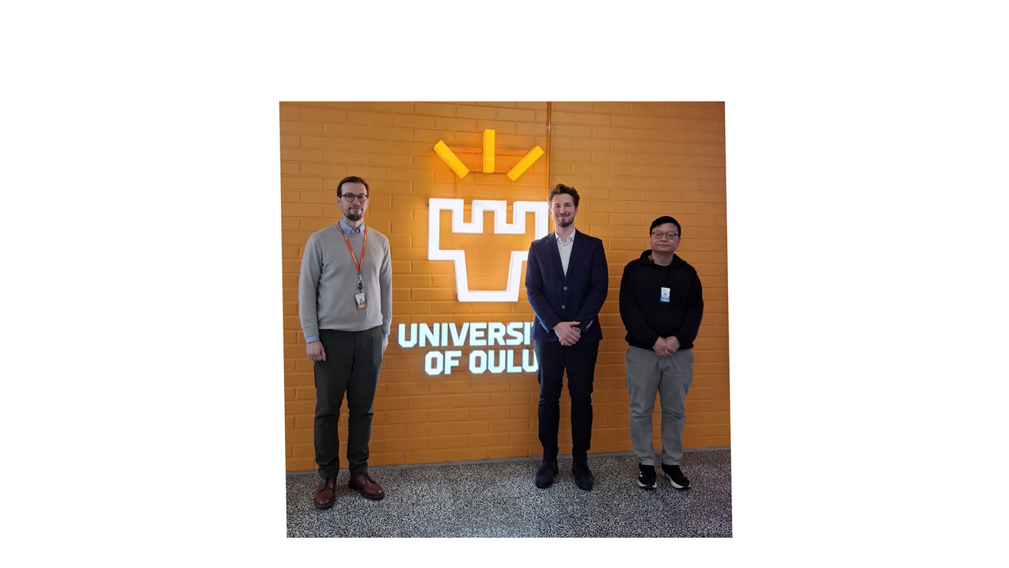 Prof. Shu, Prof. Visuri, and Kavic in front of the University of Oulu logo.