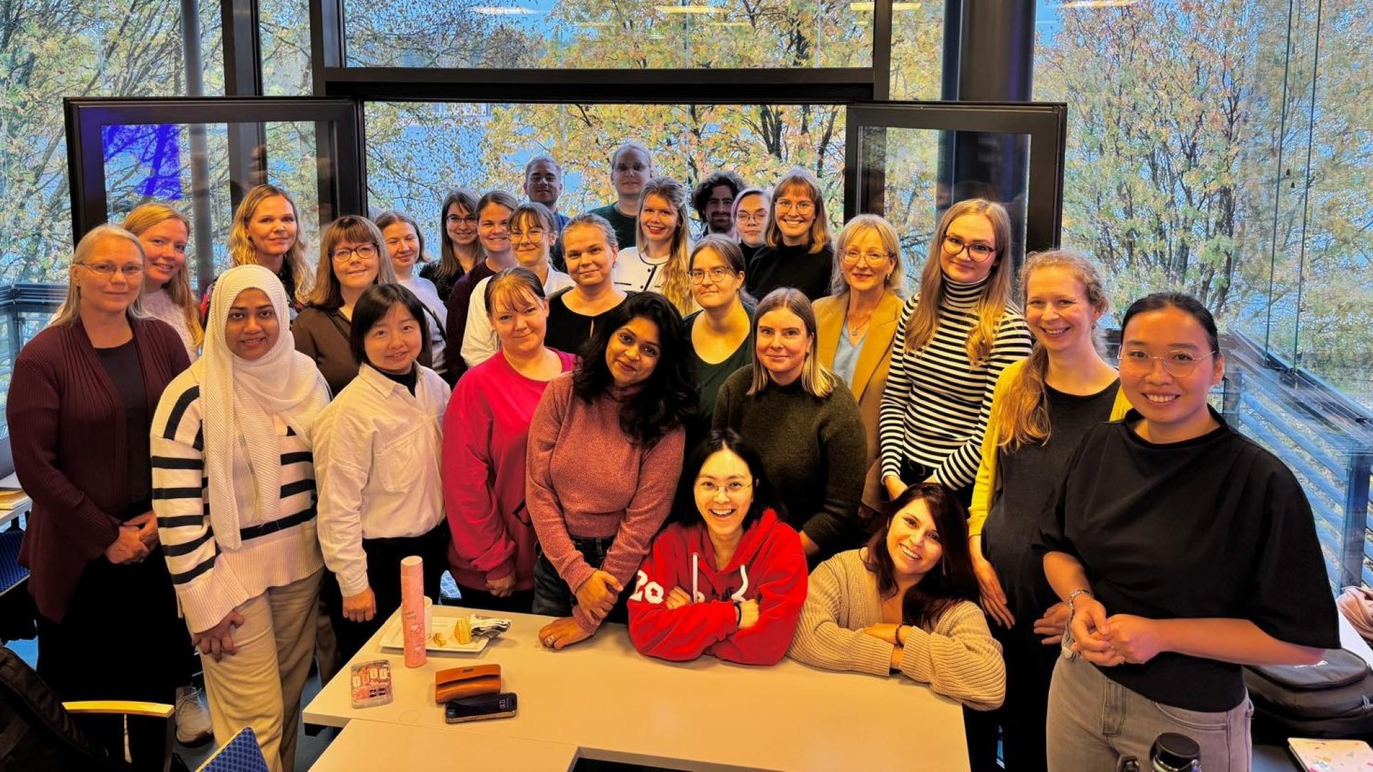 A group of smiling people standing in a classroom, with autumn trees showing in the background